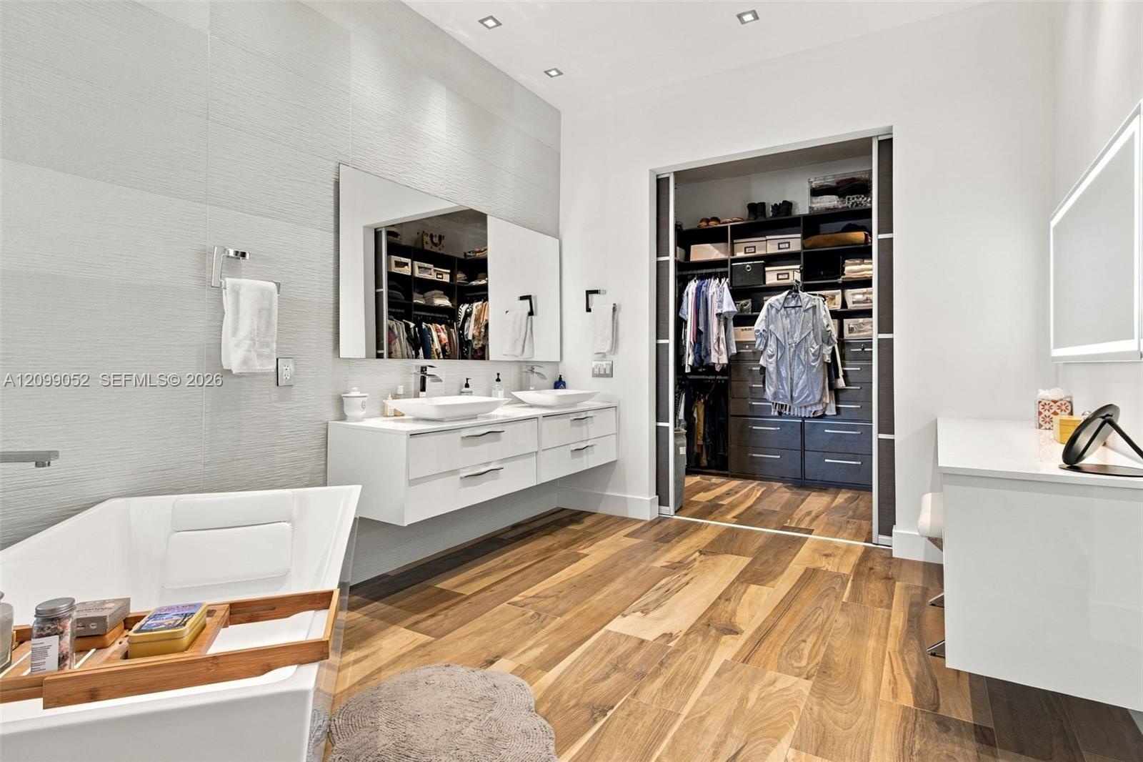 Floating white dual-sink vanity with rectangular vessel sinks, freestanding deep-soaking tub, wood-finish flooring, and built-in closet system with sl