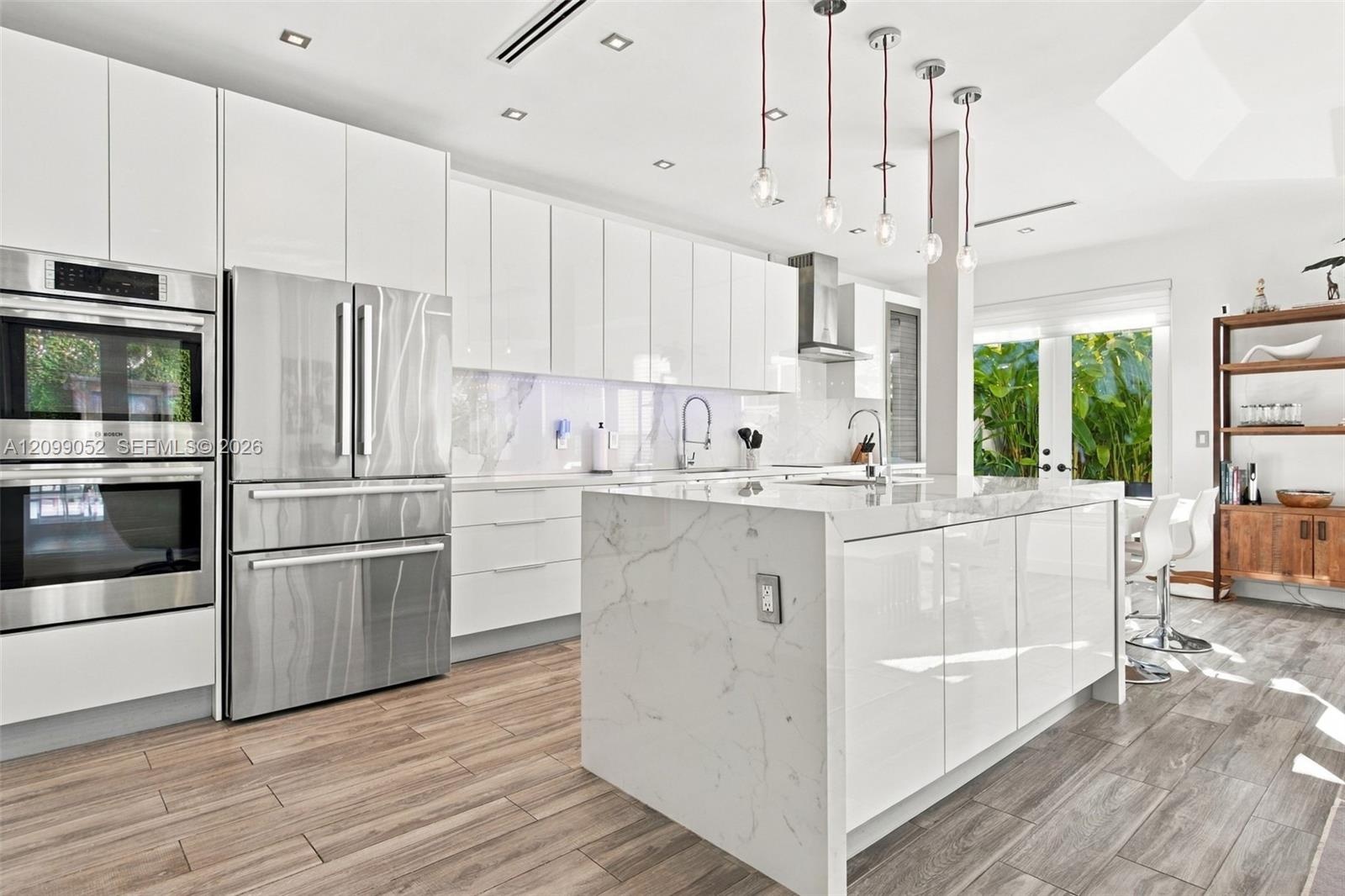 Modern kitchen featuring high-gloss white cabinetry, a center island with a stone-finish waterfall countertop, stainless steel double wall ovens, wood