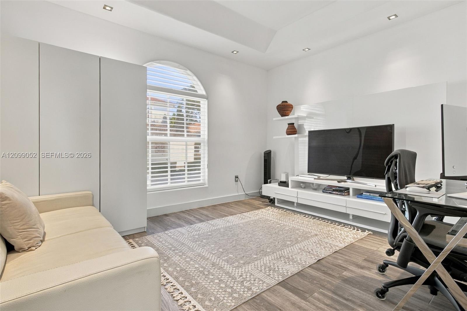 Tray ceiling with recessed lighting, arched window with horizontal blinds, built-in floor-to-ceiling cabinetry, and wood-finish flooring