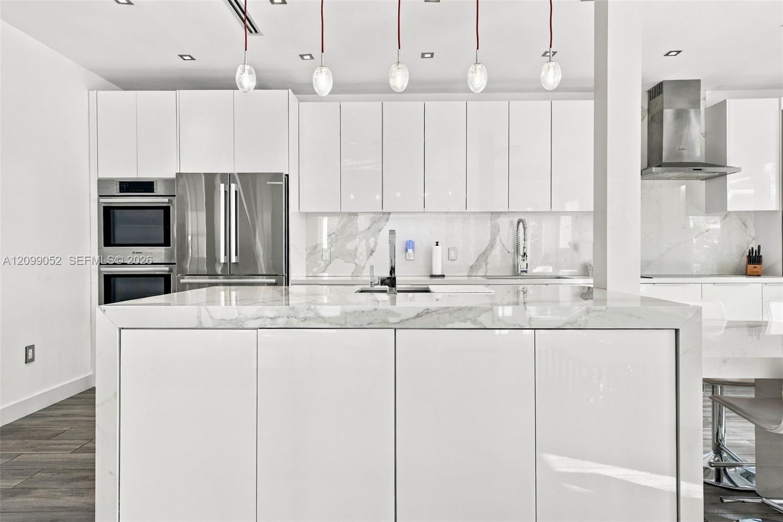 Kitchen featuring high-gloss white cabinetry, a waterfall-edge stone island, stainless steel built-in double wall ovens, a coordinating range hood, an
