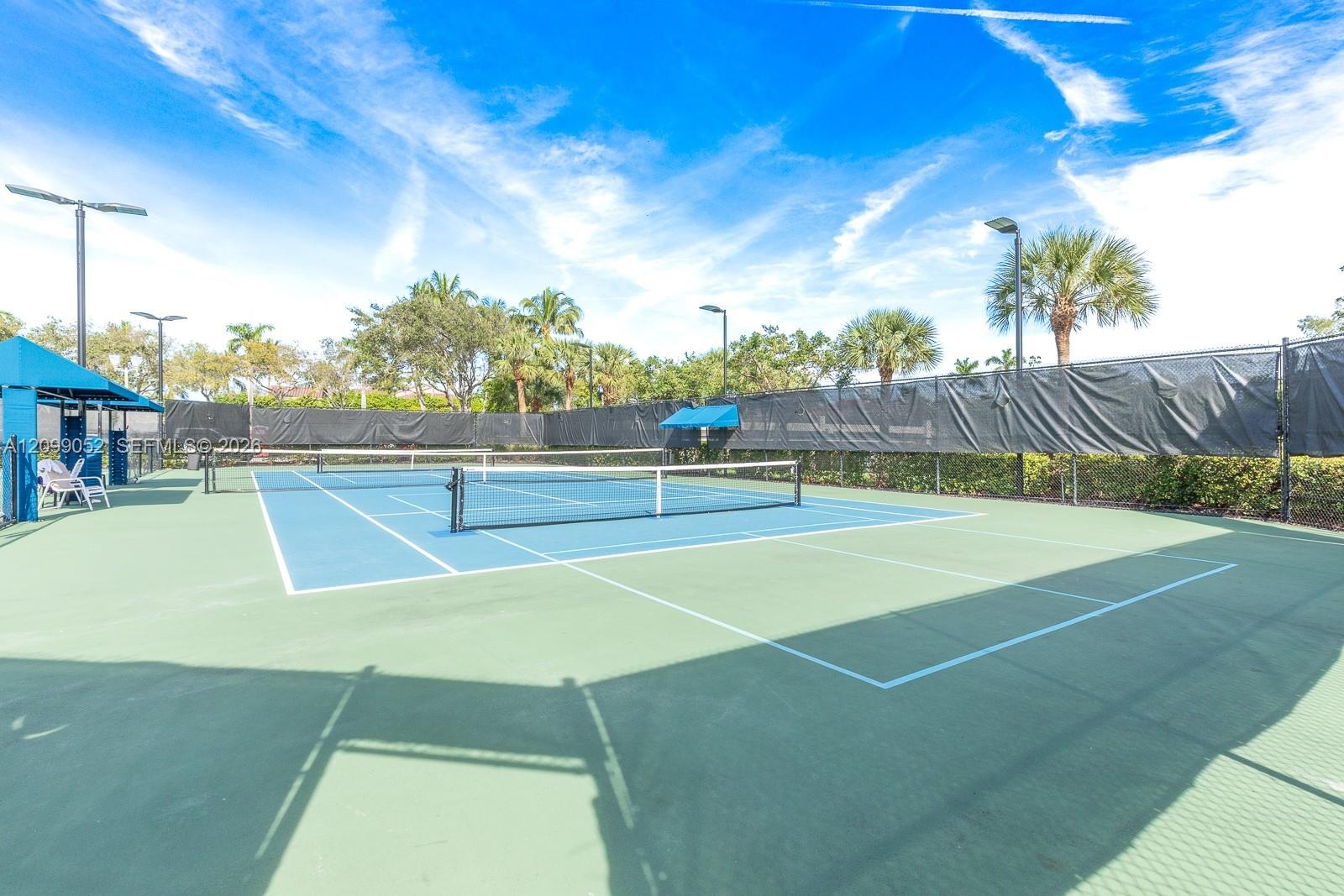 Tennis court featuring a multi-tone hard surface, perimeter fencing, overhead lighting, and shaded seating areas