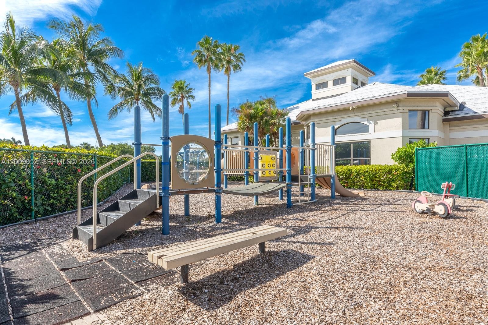 Recreational area featuring a multi-component play structure, wood-chip surface, built-in bench seating, and landscaped perimeter hedges