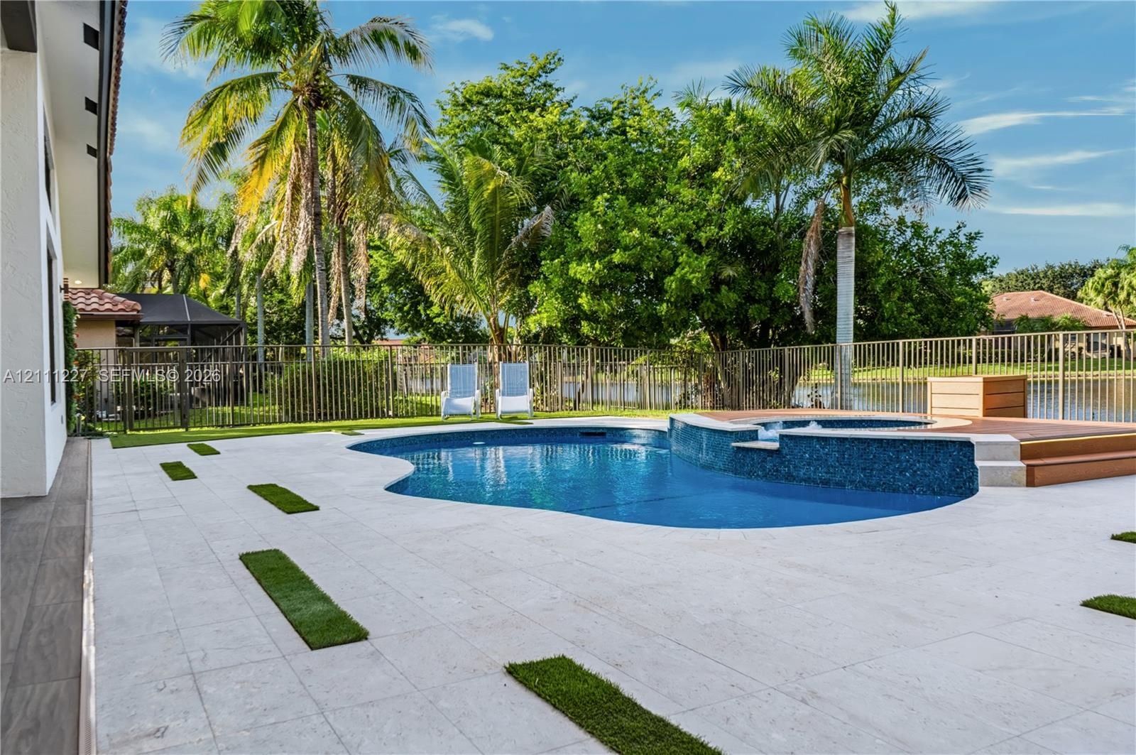 Custom-shaped swimming pool featuring a mosaic-tiled spillover spa, expansive light-toned stone patio, integrated grass accents, and a metal perimeter