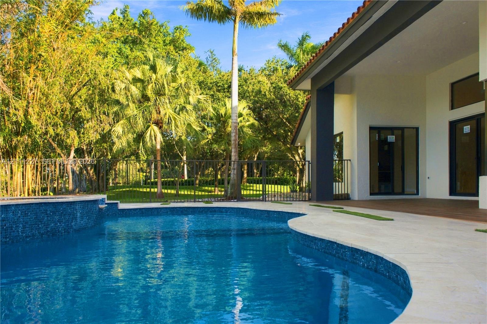 Custom swimming pool with blue tile finish, stone paver decking, covered patio, metal perimeter fencing, and mature landscaping