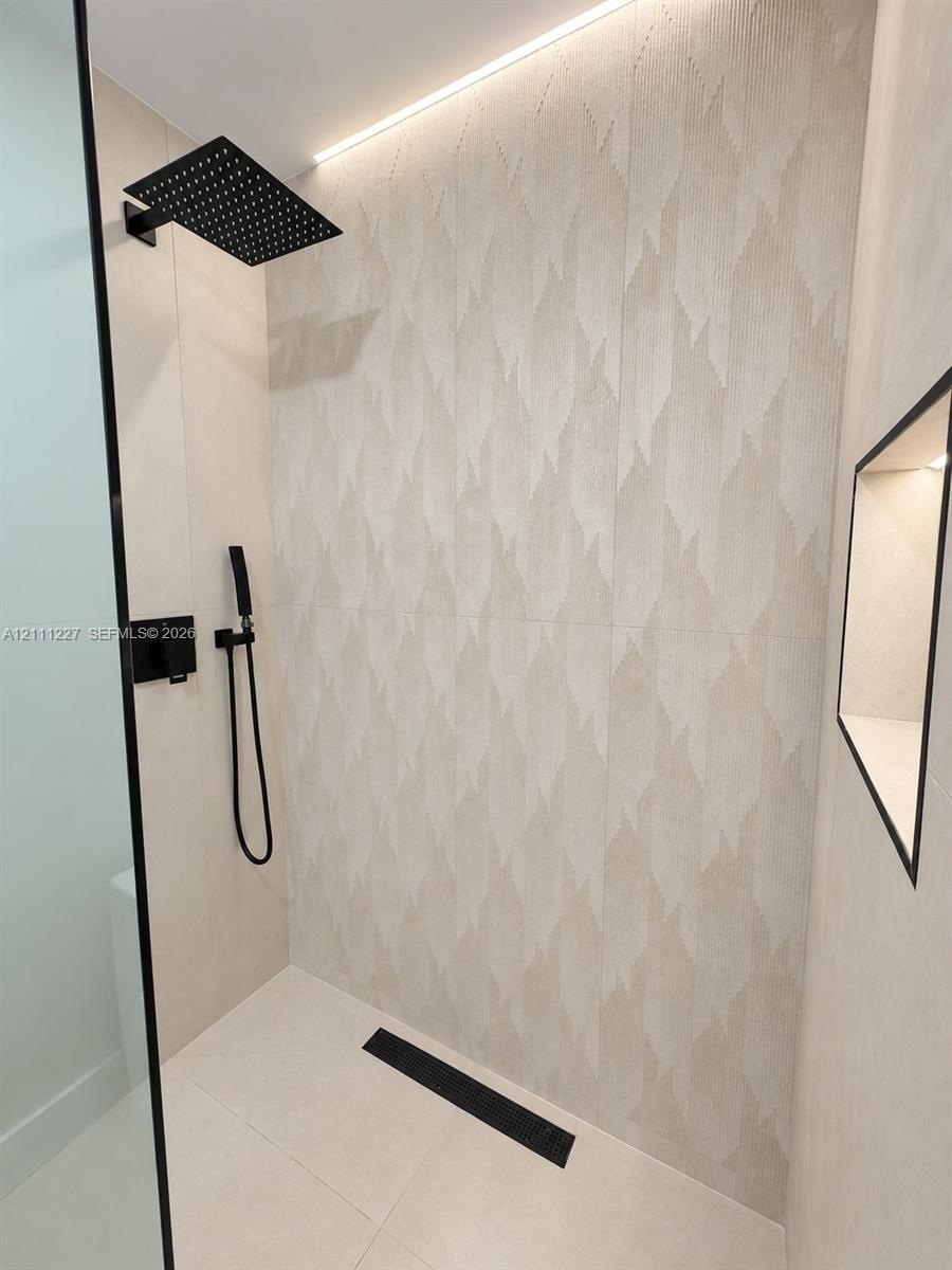 Walk-in shower featuring patterned large-format wall tile, matte black rainfall and handheld showerheads, integrated recessed niche lighting, and a li
