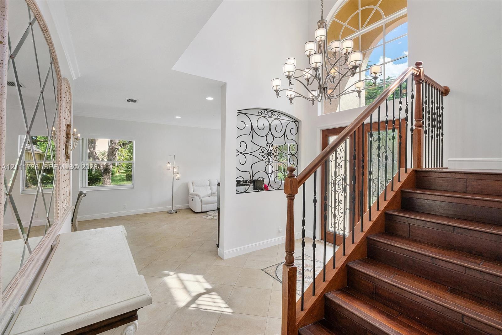 Grand foyer featuring a high ceiling, wood-finish staircase with decorative metal balusters, arched window, wrought-iron wall accent, and multi-arm ch