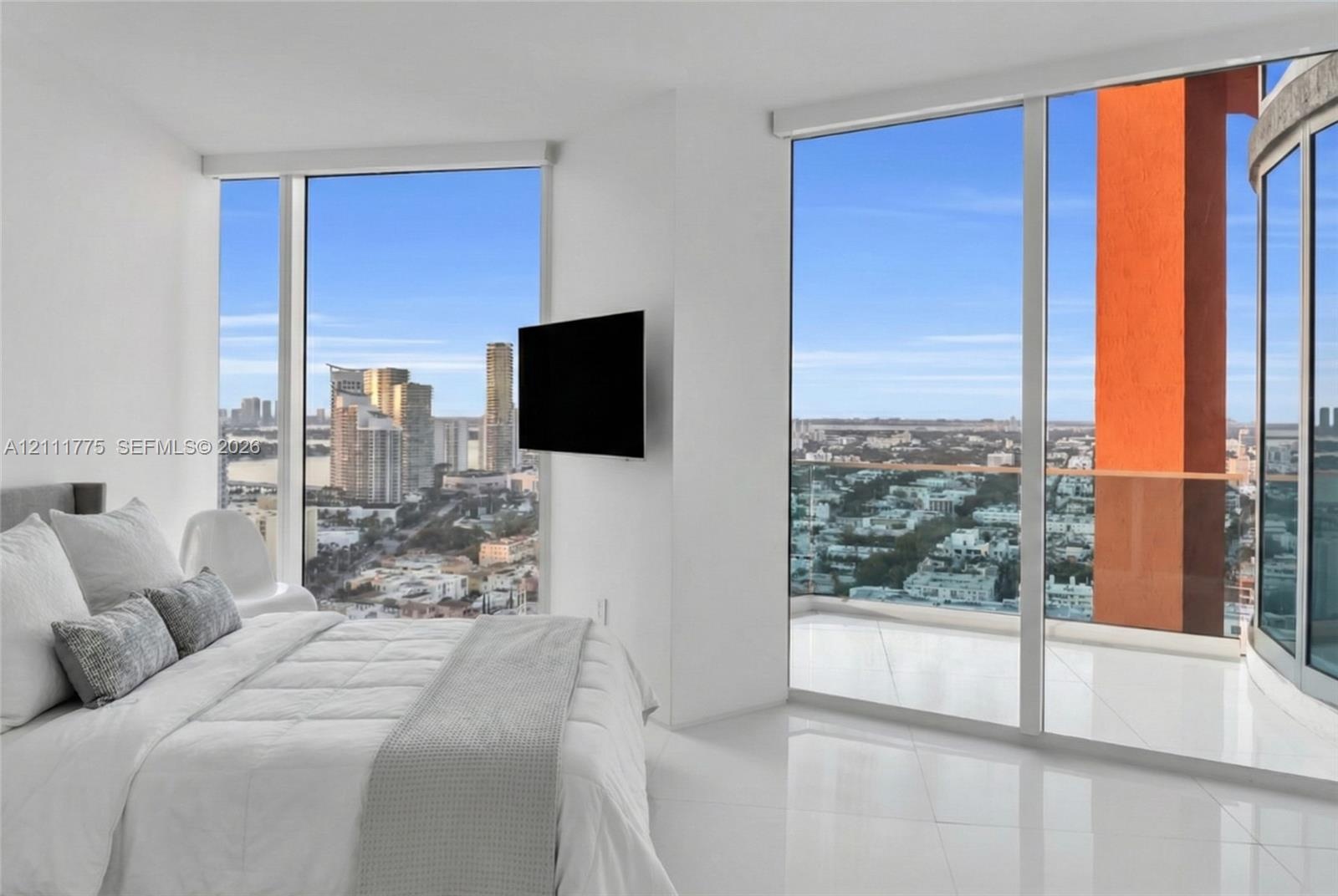 Floor-to-ceiling windows, white tile flooring, private balcony with glass railing, and wall-mounted television hardware