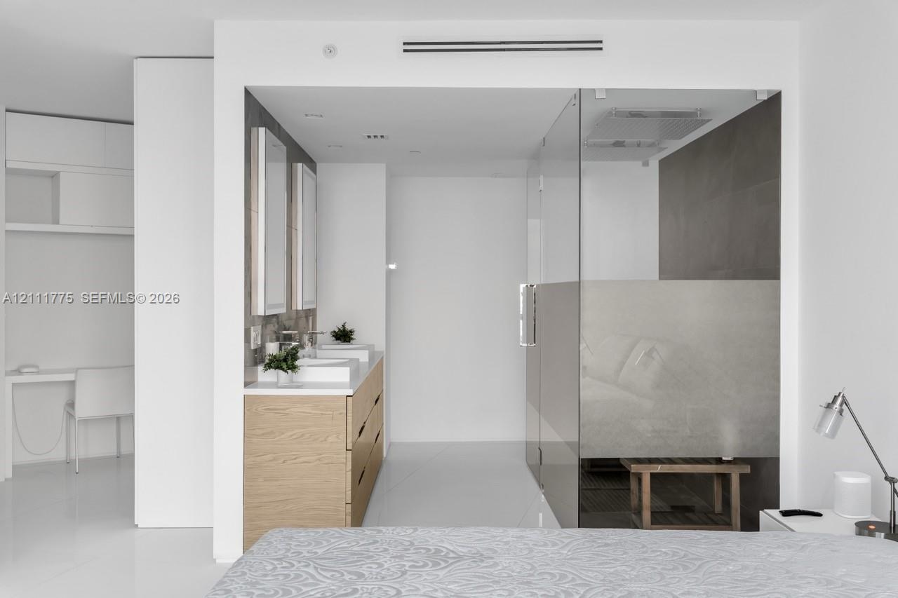 Wood-finish double vanity with rectangular sinks, white countertop, and frameless glass-enclosed shower with large-format tile wall