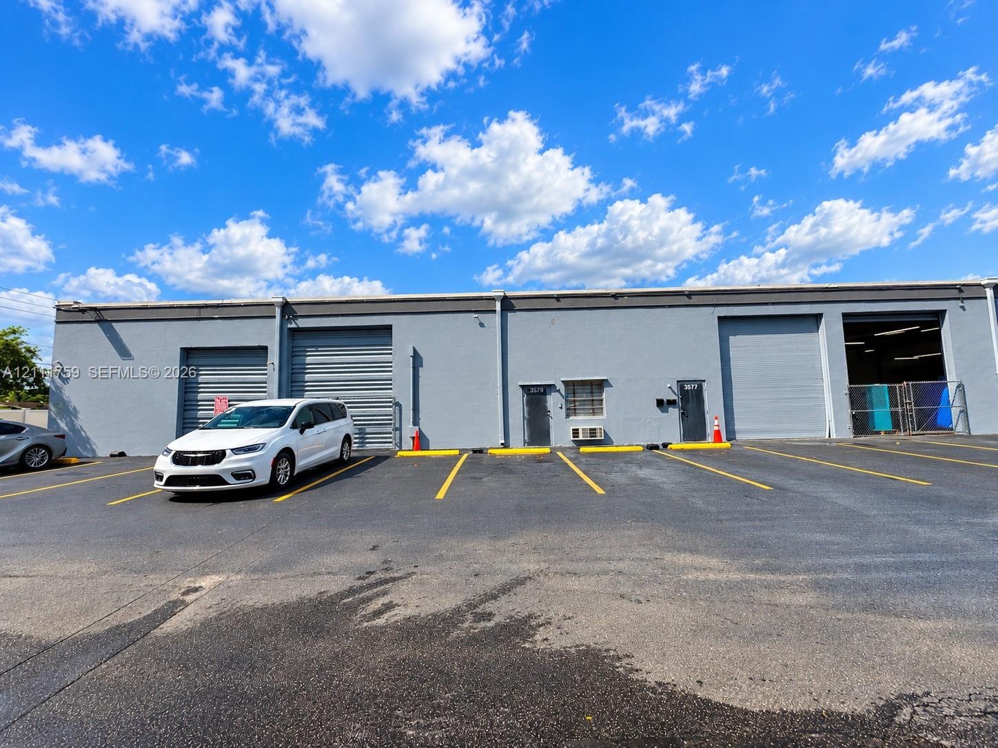 Single-story industrial exterior featuring a smooth gray concrete facade, three metal roll-up doors, two dedicated entry points, and an asphalt-paved 