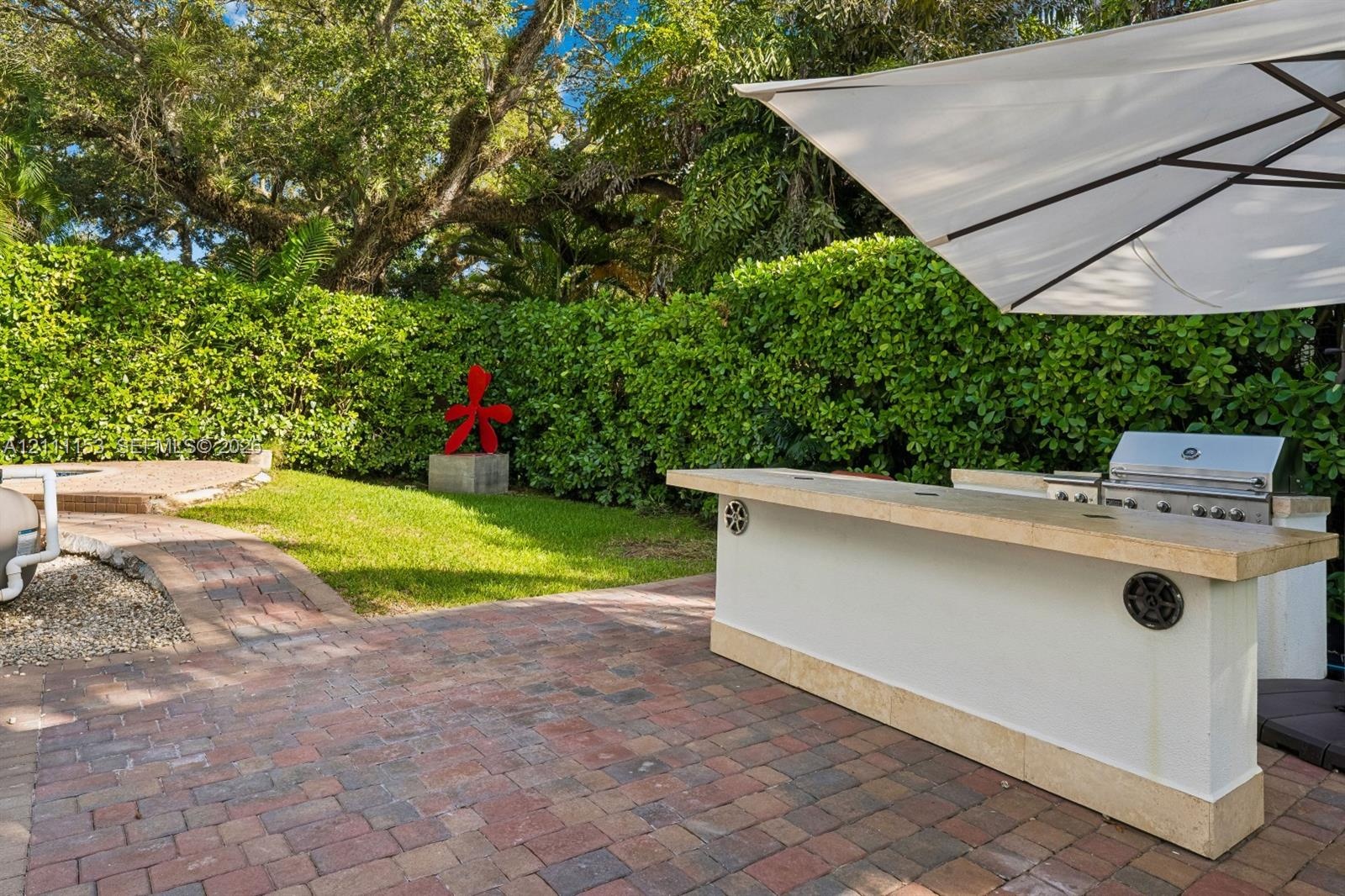 Outdoor kitchen featuring a built-in grill and stone-capped bar, set upon a red brick paver patio, surrounded by mature privacy hedging and a manicure