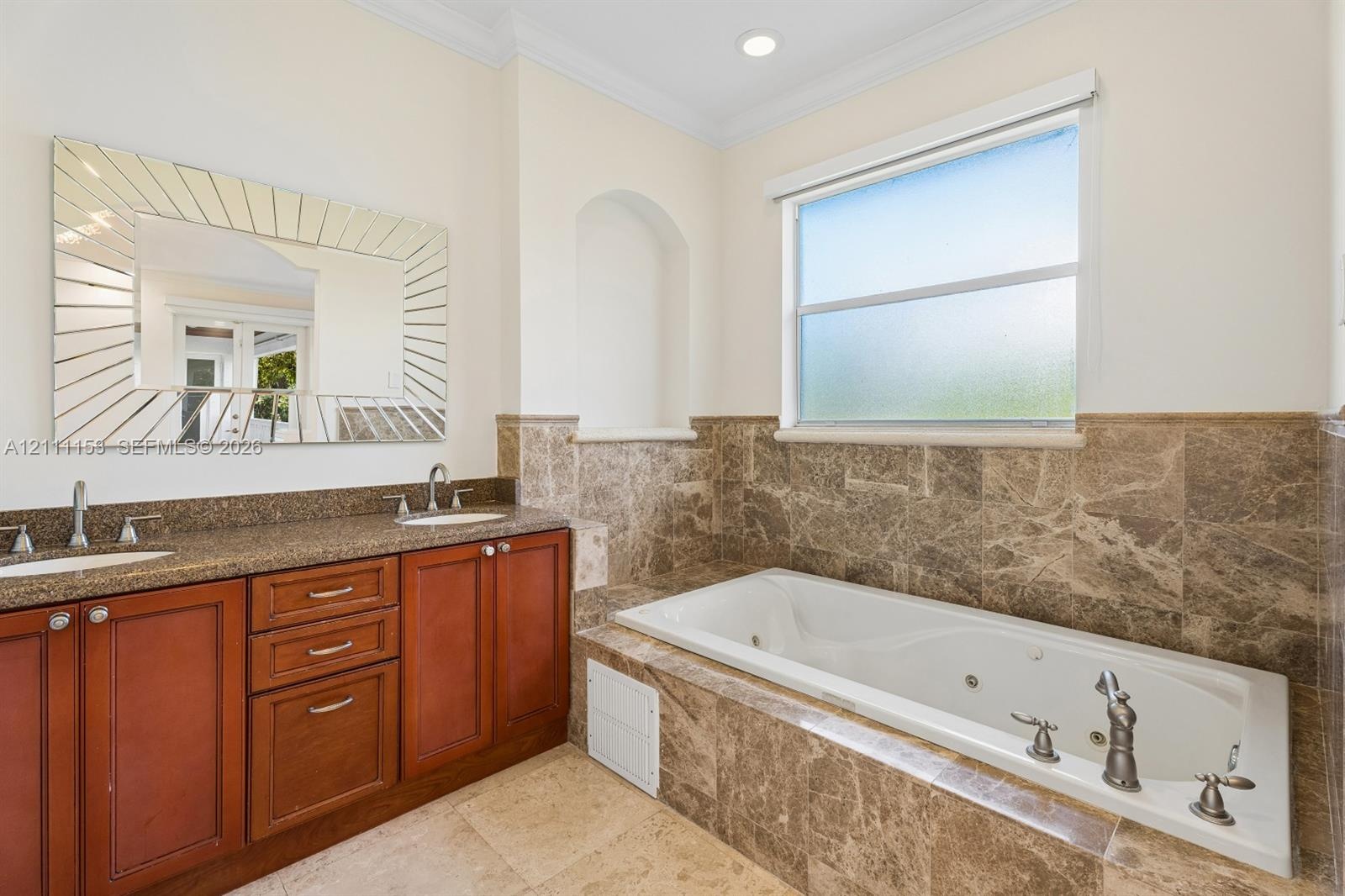 Dual-sink vanity with stone countertop, soaking tub with jetted features, full-height stone tile surround, built-in recessed wall niche, and decorativ