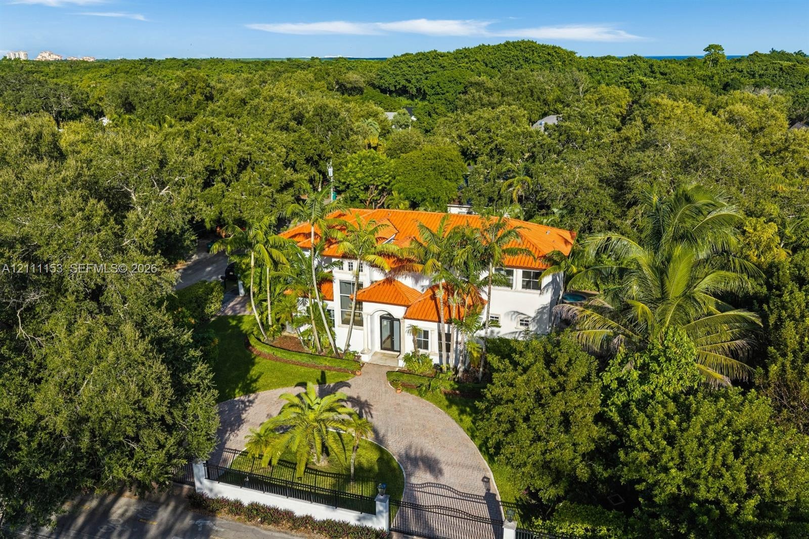Two-story residence featuring a terracotta-tiled roof, white exterior, arched entryway, paved circular driveway, and mature palm trees