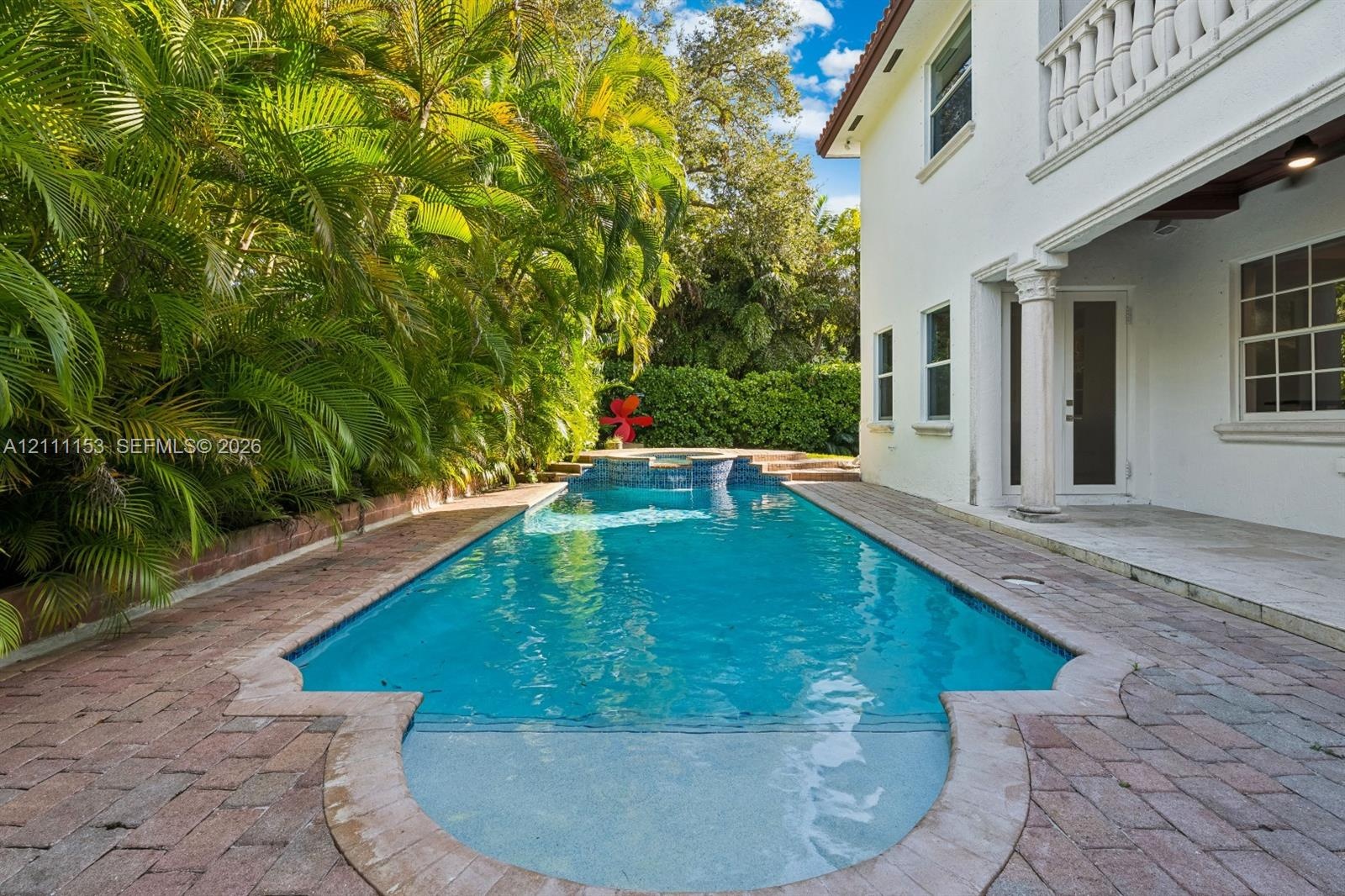 Rectangular pool featuring a built-in spa, integrated sun shelf, expansive brick paver decking, and a covered patio area with a structural column