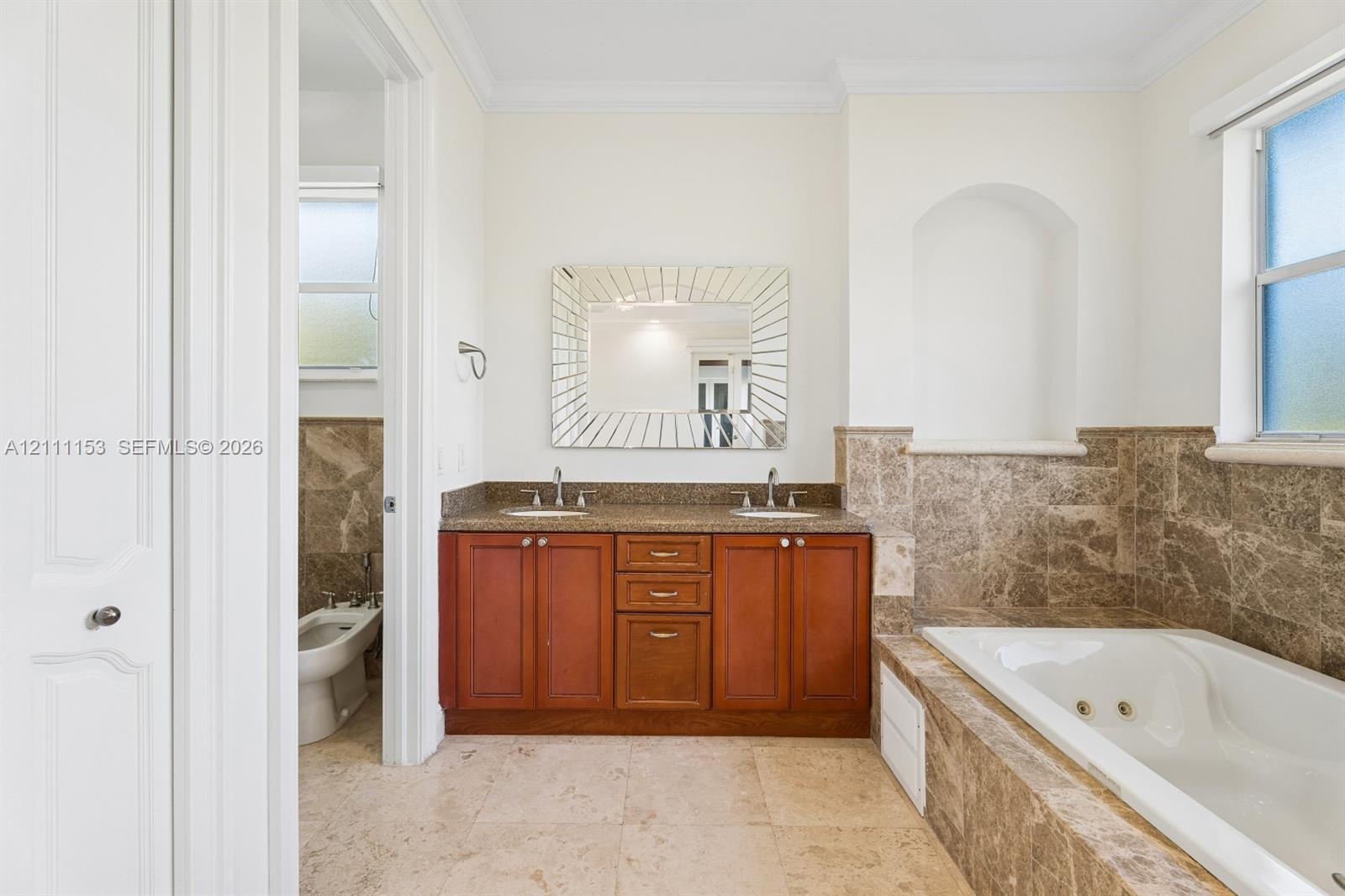 Dual-sink vanity with wood-finish cabinetry, natural stone countertop, recessed wall niche, tiled soaking tub, and stone-finish flooring