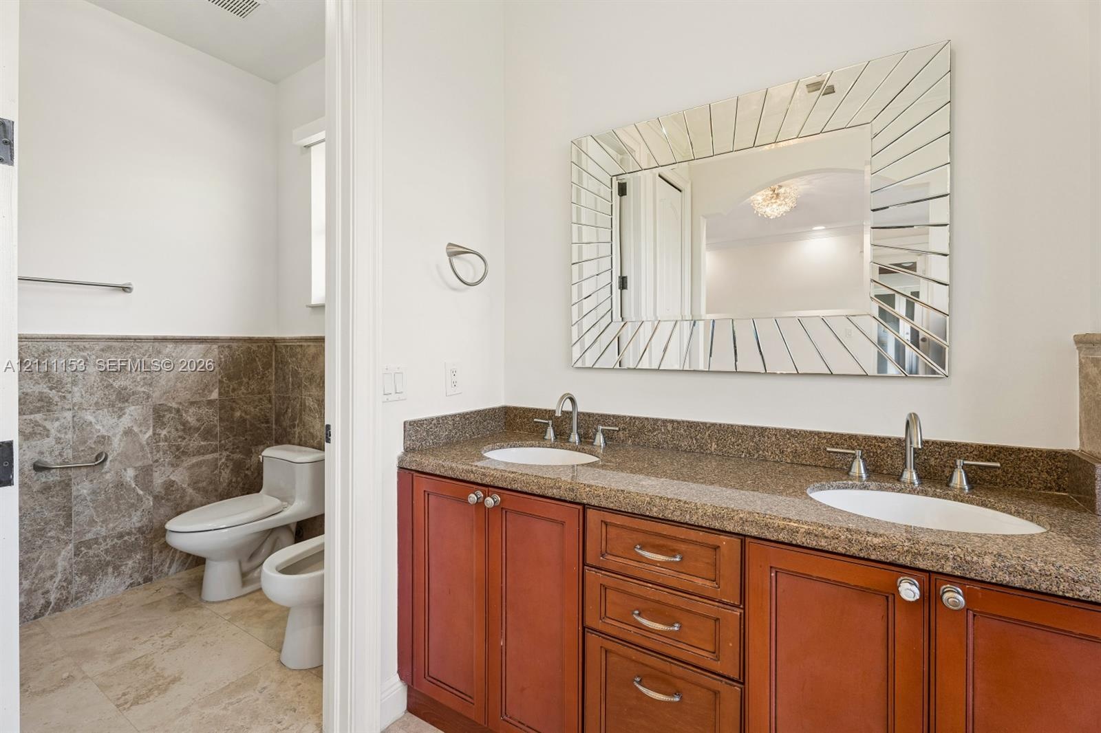 Dual-sink vanity with stone countertop and cabinetry, decorative sunburst-style wall mirror, stone tile wainscoting, and ceramic flooring