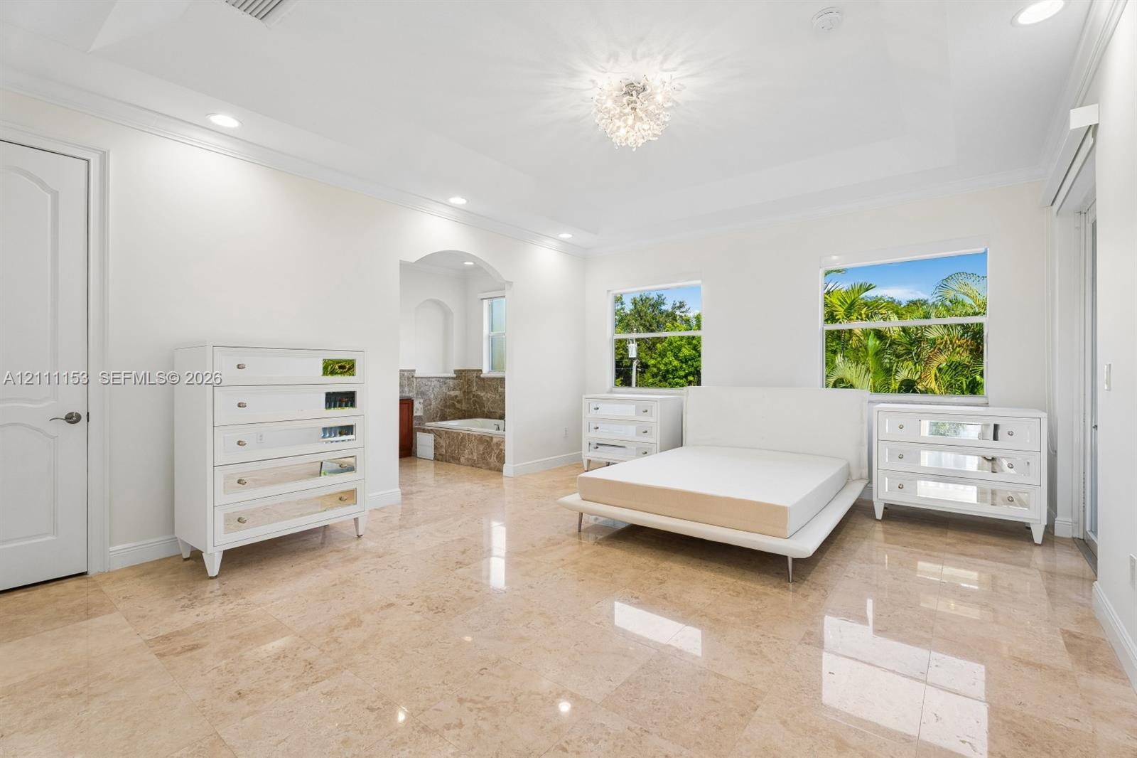 Tray ceiling with recessed lighting, central crystal-style chandelier, polished stone flooring, and arched entryway featuring a built-in tiled tub sur