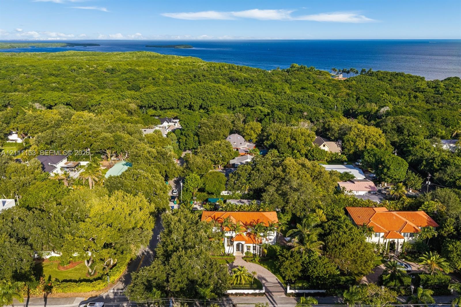 Aerial perspective featuring residences with terracotta-style roof tiles, expansive tree canopy, and proximity to the coastline