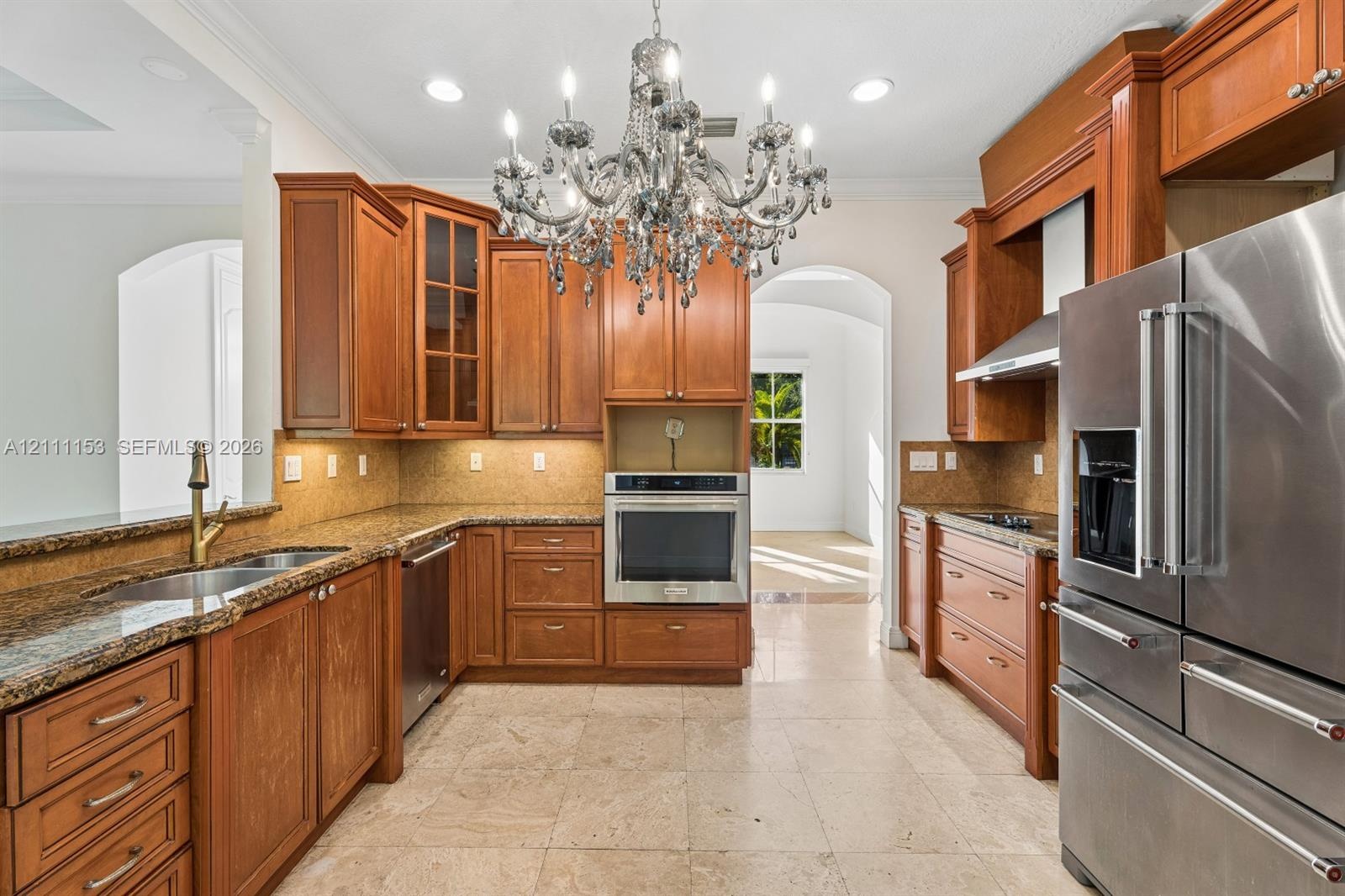 Kitchen featuring custom wood cabinetry, stone-surface countertops, built-in stainless steel appliances, a multi-tiered crystal chandelier, and large-