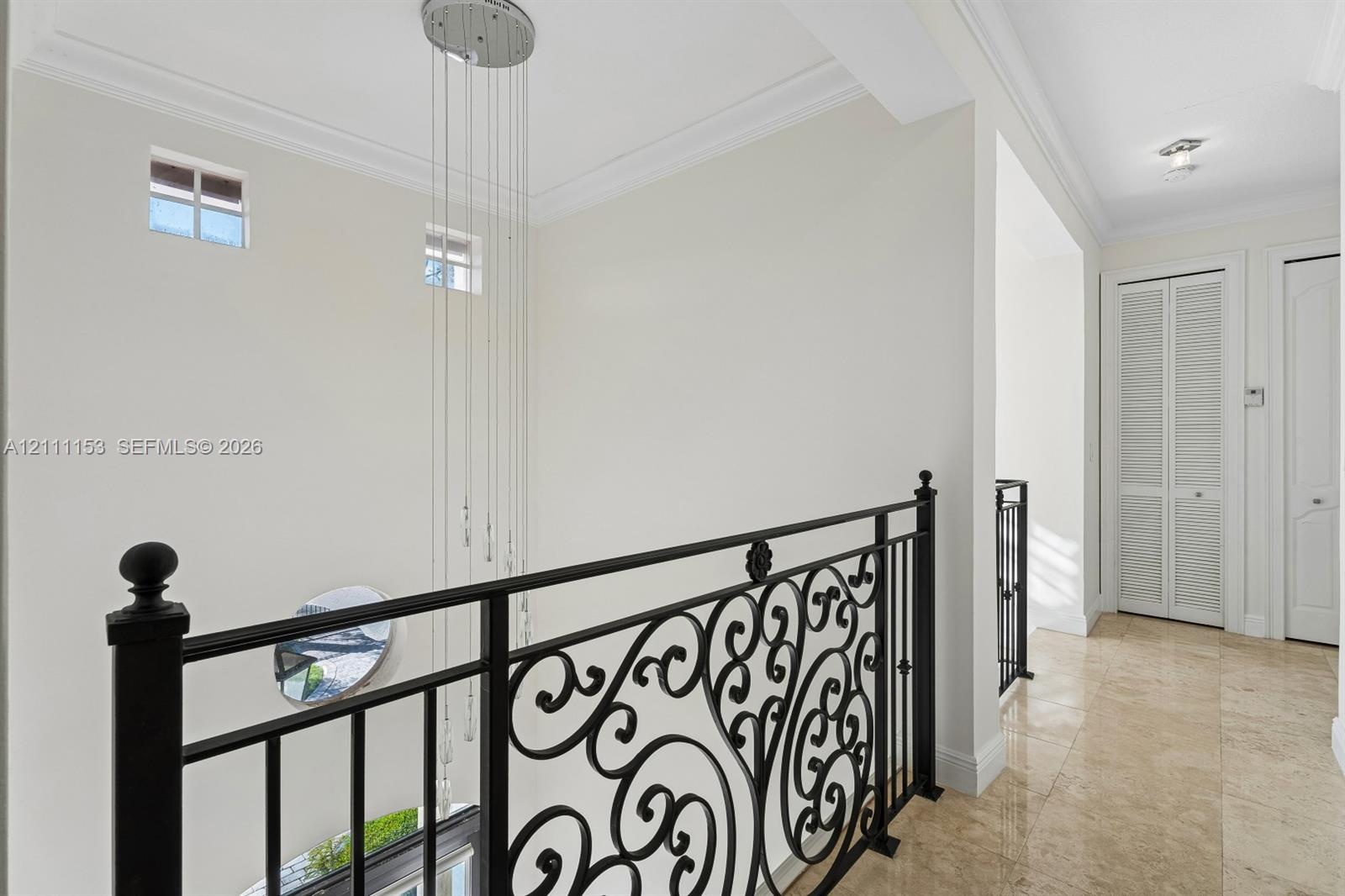 Hallway featuring polished stone flooring, intricate black iron railing, multi-tiered pendant lighting, crown molding, and louvered closet doors