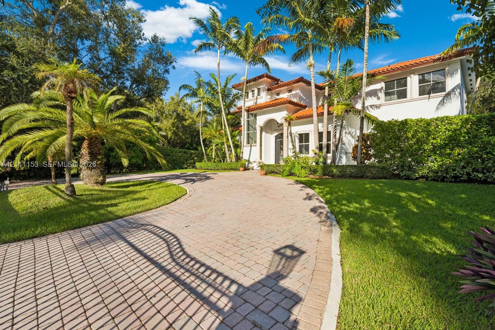 Stucco exterior featuring a terracotta tile roof, arched entryway, paved driveway, and mature palm landscaping