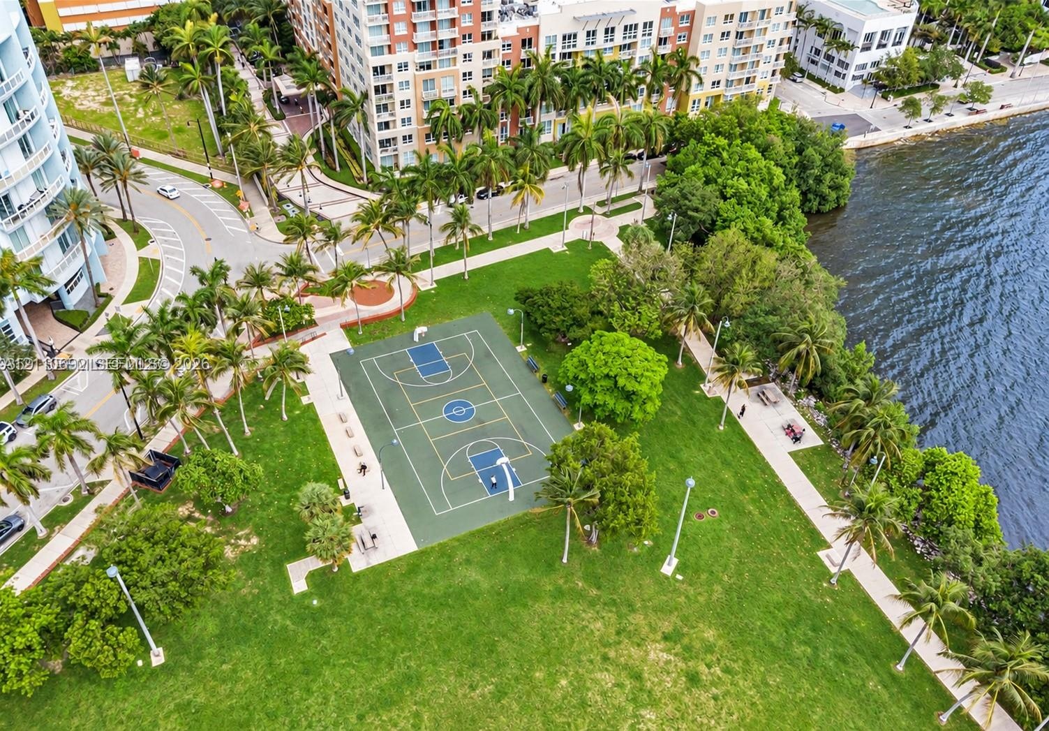 Waterfront outdoor space featuring a paved basketball court, manicured lawn, established palm trees, and pedestrian walkways