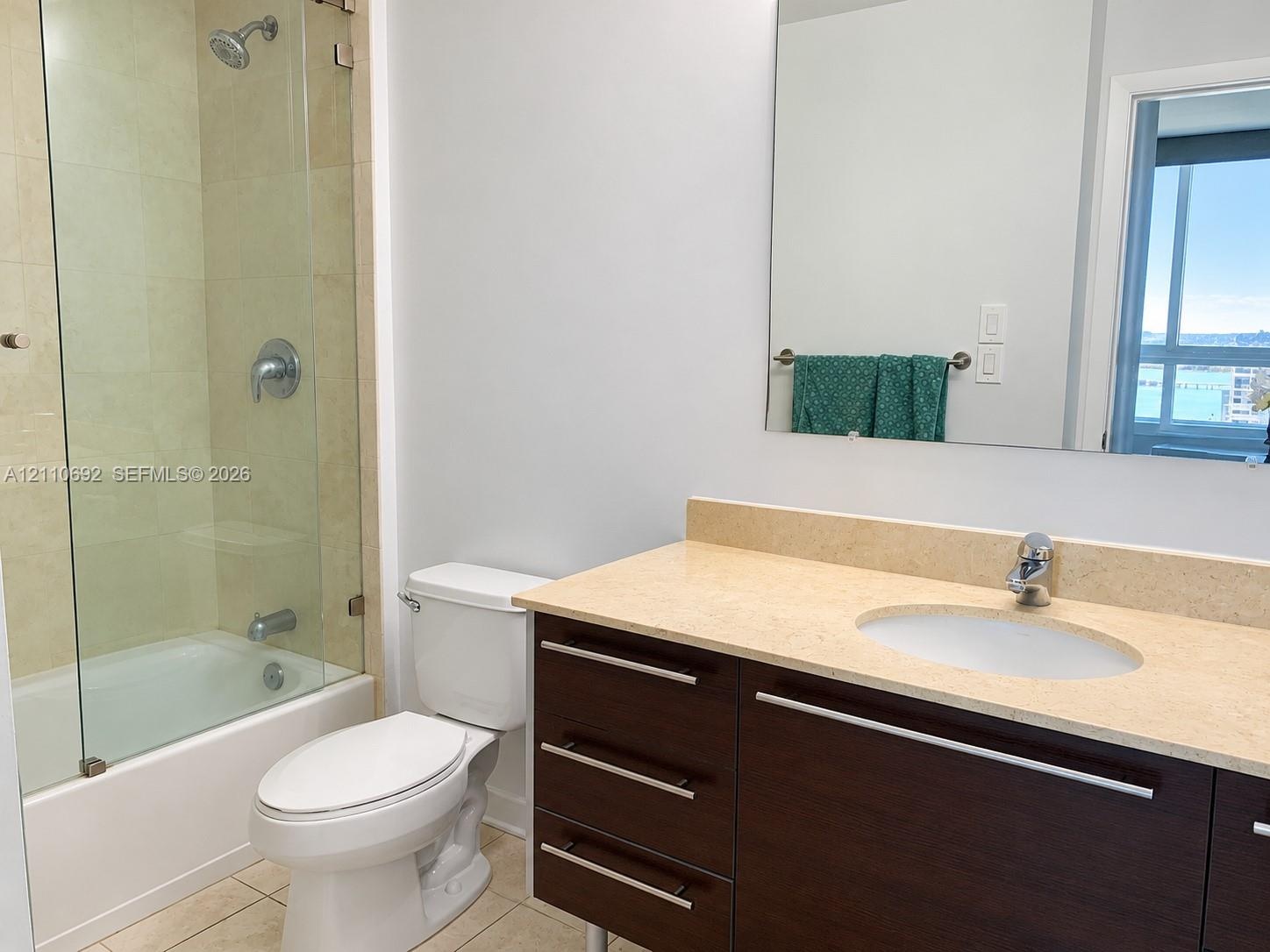 Bathroom featuring a dark wood-finish vanity with a light-toned countertop, integrated oval sink, glass-enclosed shower-tub combination with tile surr