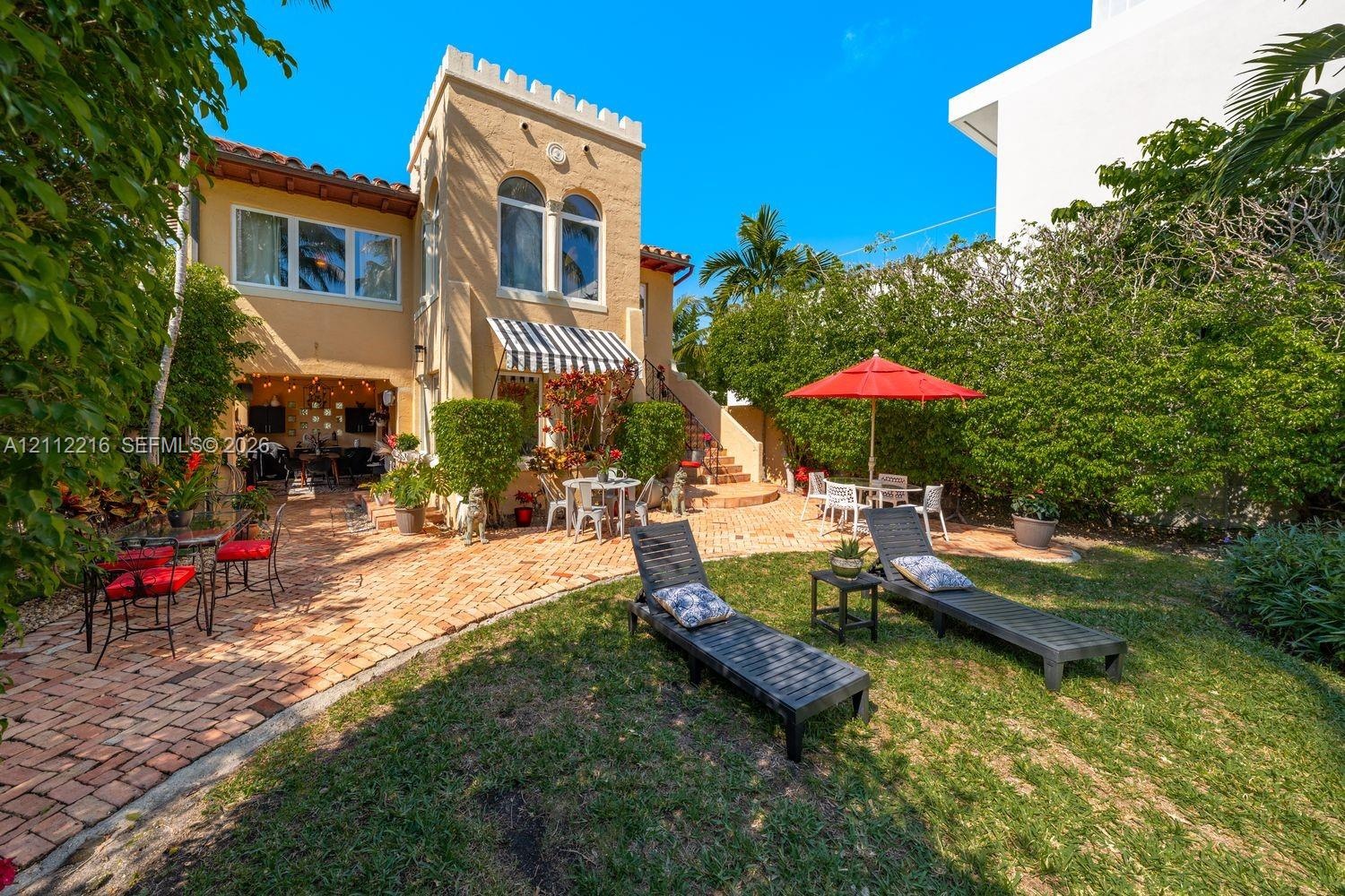 Exterior featuring a castellated tower, arched windows, and a paved brick patio area bordered by mature shrubbery and lawn