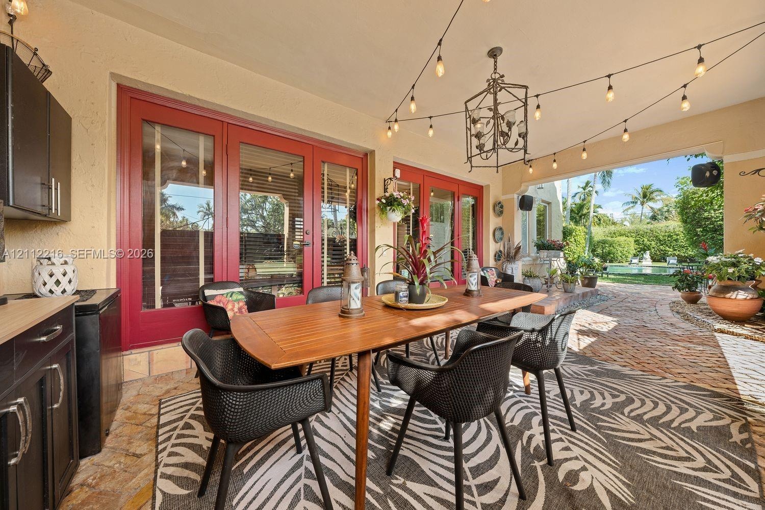 Covered patio featuring stone paver flooring, outdoor kitchen cabinetry, red-framed glass double doors, an overhead iron chandelier, and string lighti