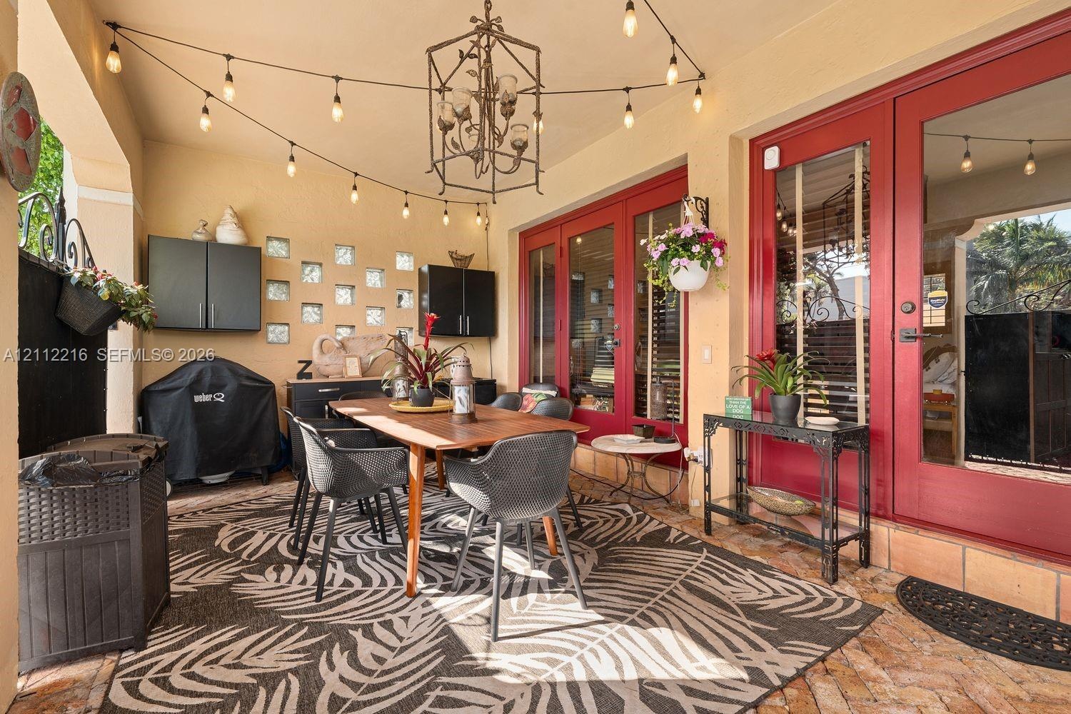 Covered patio featuring brick flooring, dual glass-paned doors with bold red frames, decorative glass block accents, and an overhead geometric chandel