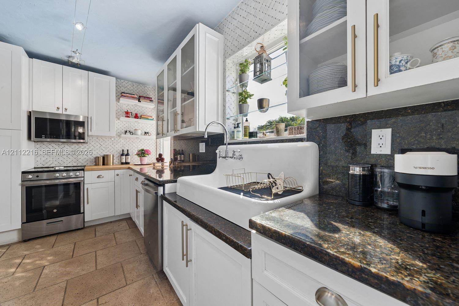 Kitchen featuring white cabinetry with glass-front uppers, apron-front farmhouse sink, stone countertops, stainless steel appliances, and patterned ti