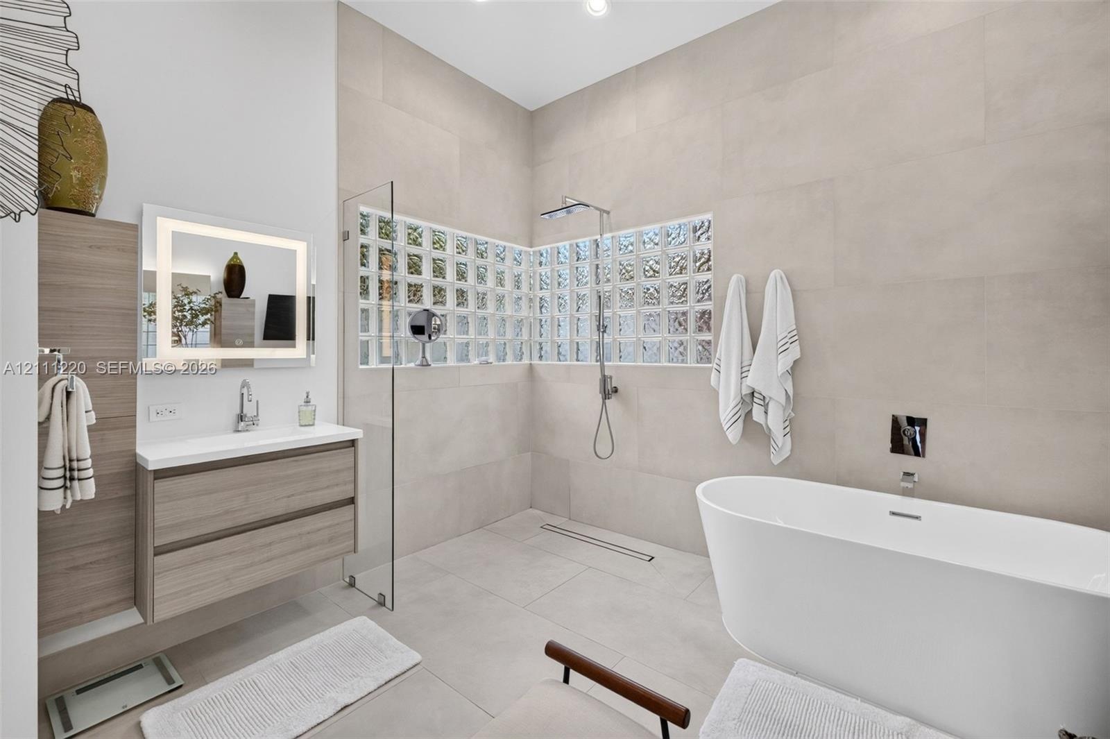 Bathroom featuring a freestanding soaking tub, walk-in shower with glass partition, neutral large-format wall tile, wood-finish wall-mounted vanity, a