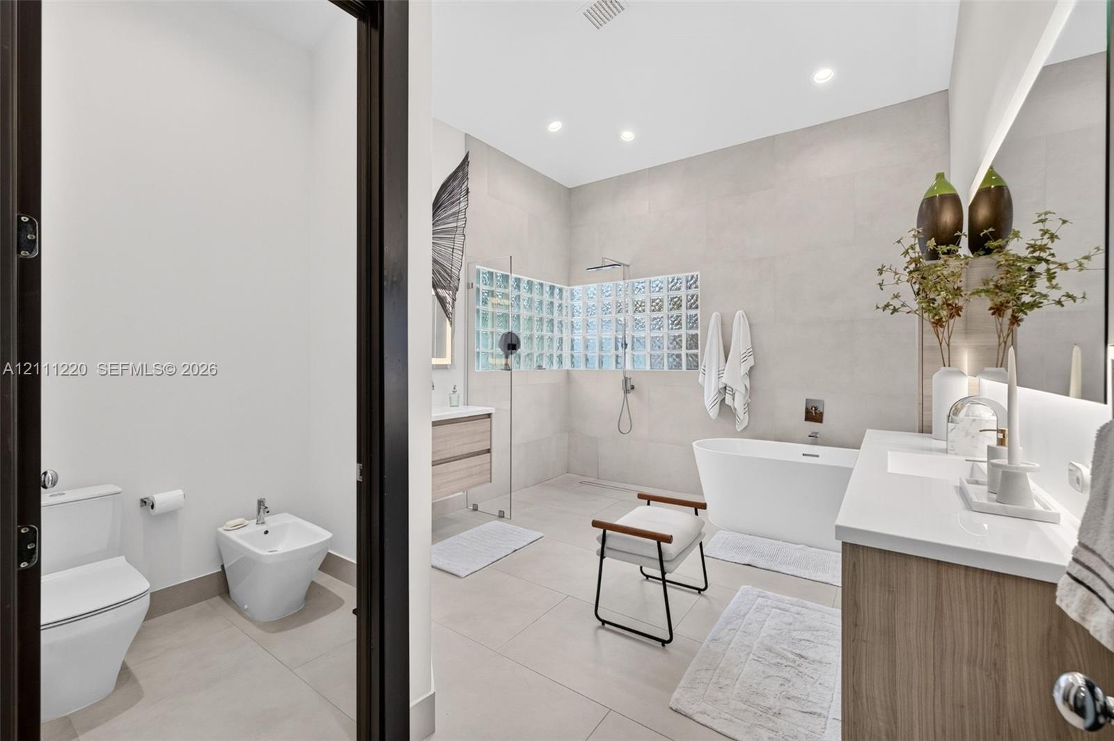 Bathroom featuring a large freestanding soaking tub, walk-in shower with glass block accent wall, double vanity with wood-finish cabinetry, wall-mount