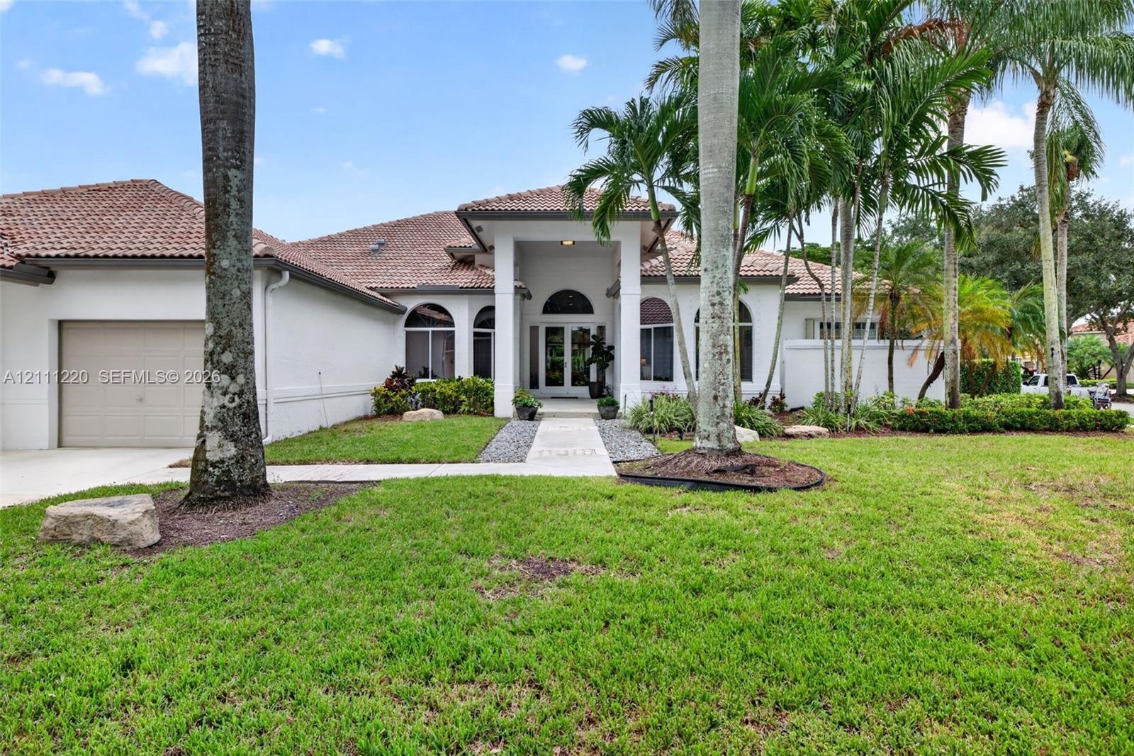 Stucco exterior featuring a tile roof, arched windows, covered entryway, and manicured landscaping with mature palm trees