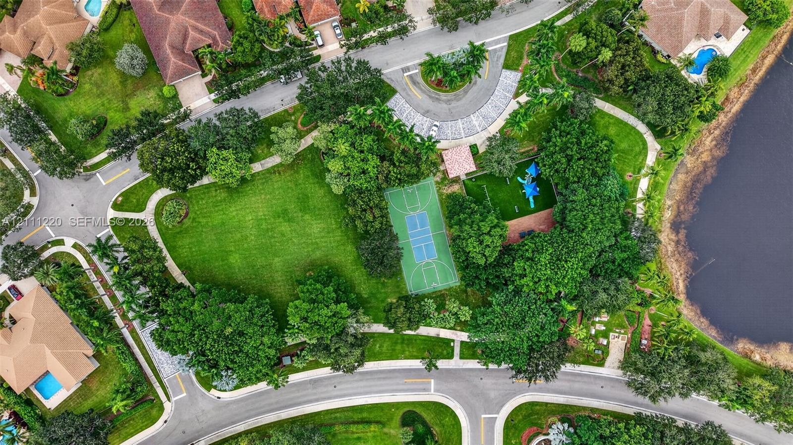 Aerial perspective of a community recreation area featuring a basketball court, playground, expansive green space, mature trees, and a bordering water