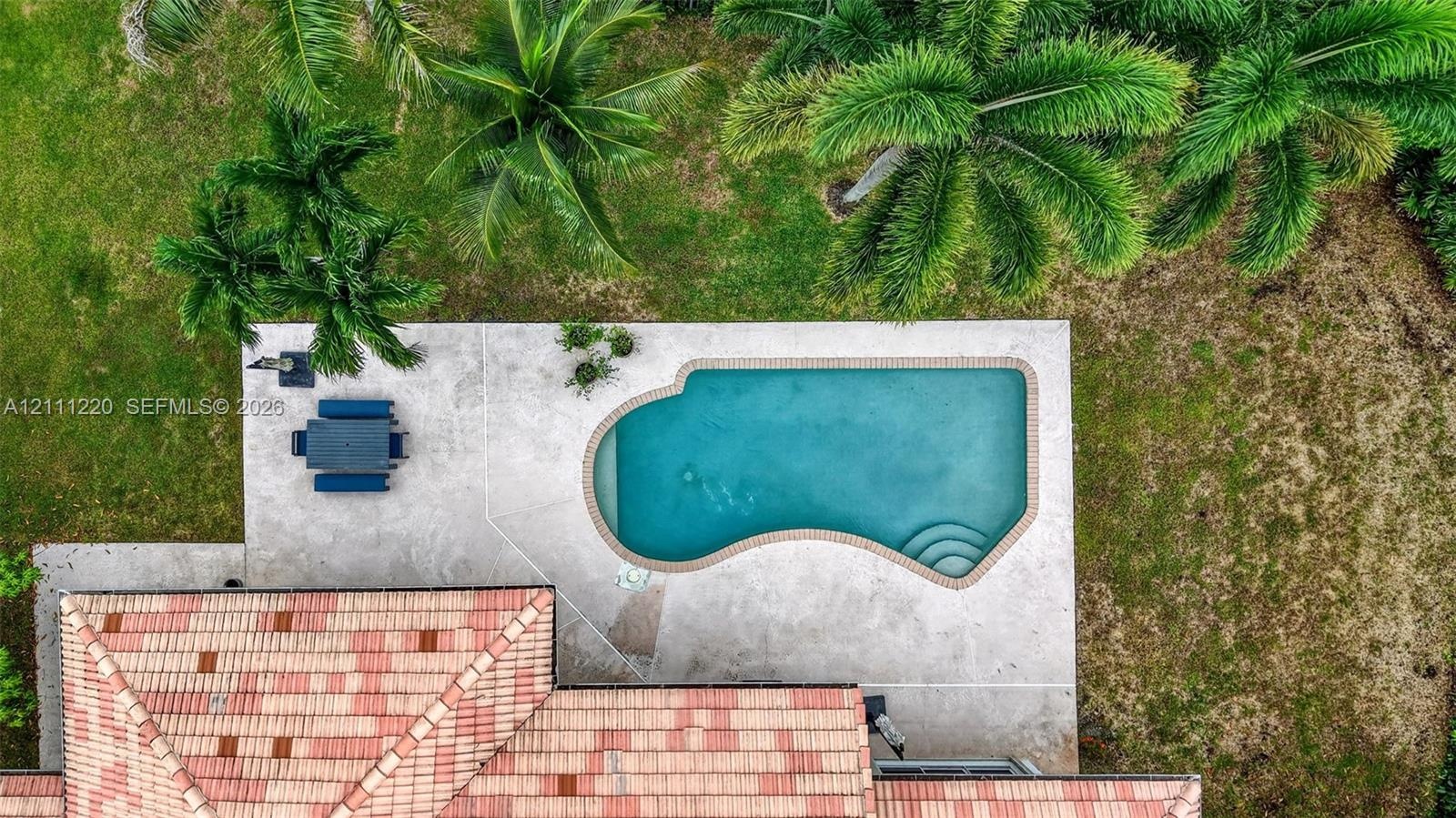 Aerial view of a sprawling property featuring an expansive concrete pool deck, in-ground swimming pool with integrated steps, terra-cotta tile roofing