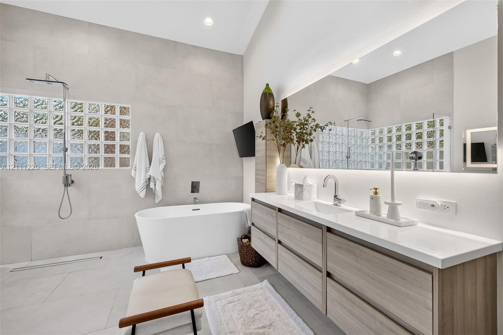 Bathroom featuring a floating wood-finish vanity with a countertop basin, a standalone soaking tub, full-wall tiling, a glass-block privacy window, an