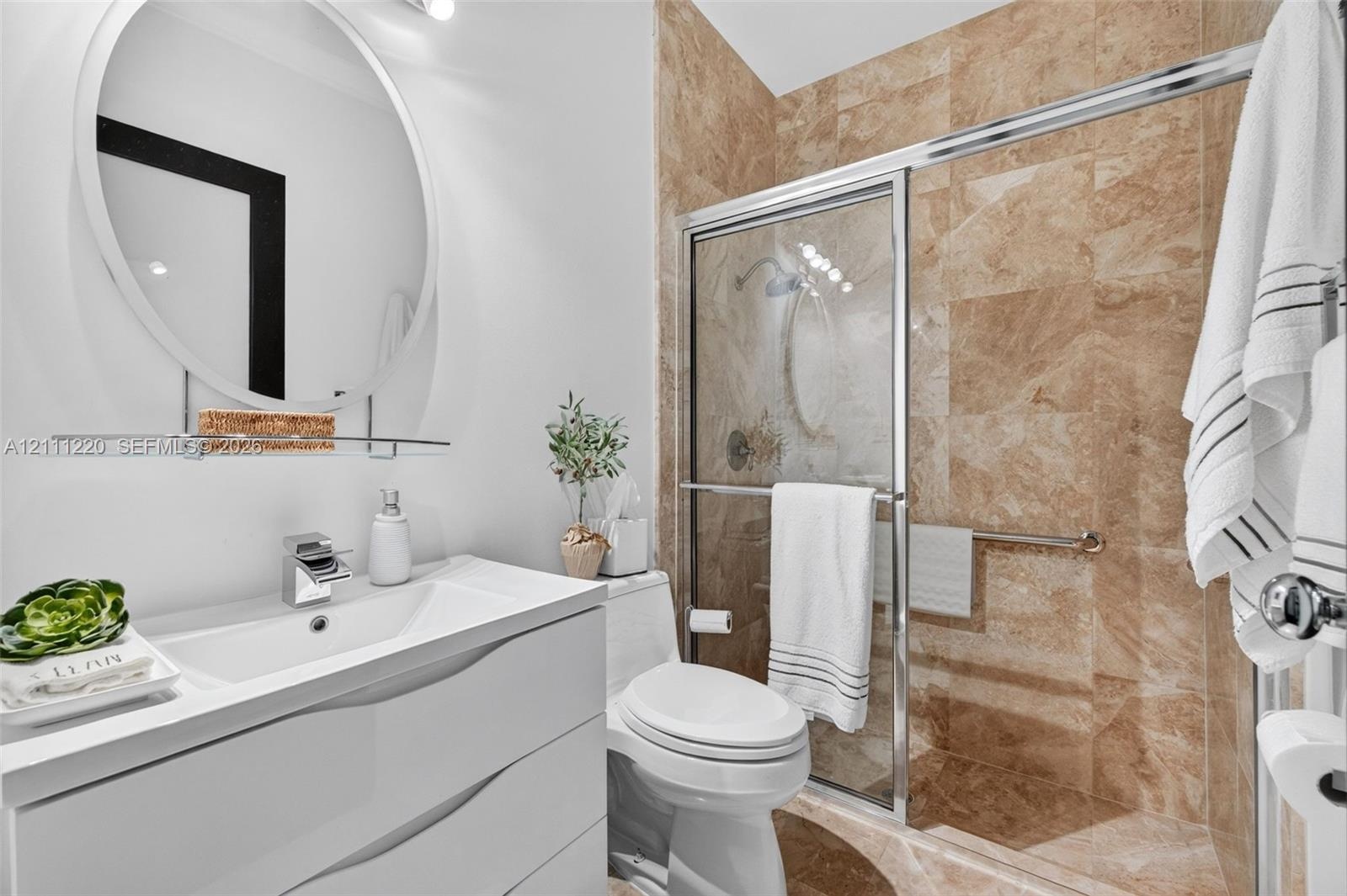 Contemporary white vanity with integrated basin, polished chrome faucet, circular wall mirror, and walk-in shower featuring floor-to-ceiling stone-loo