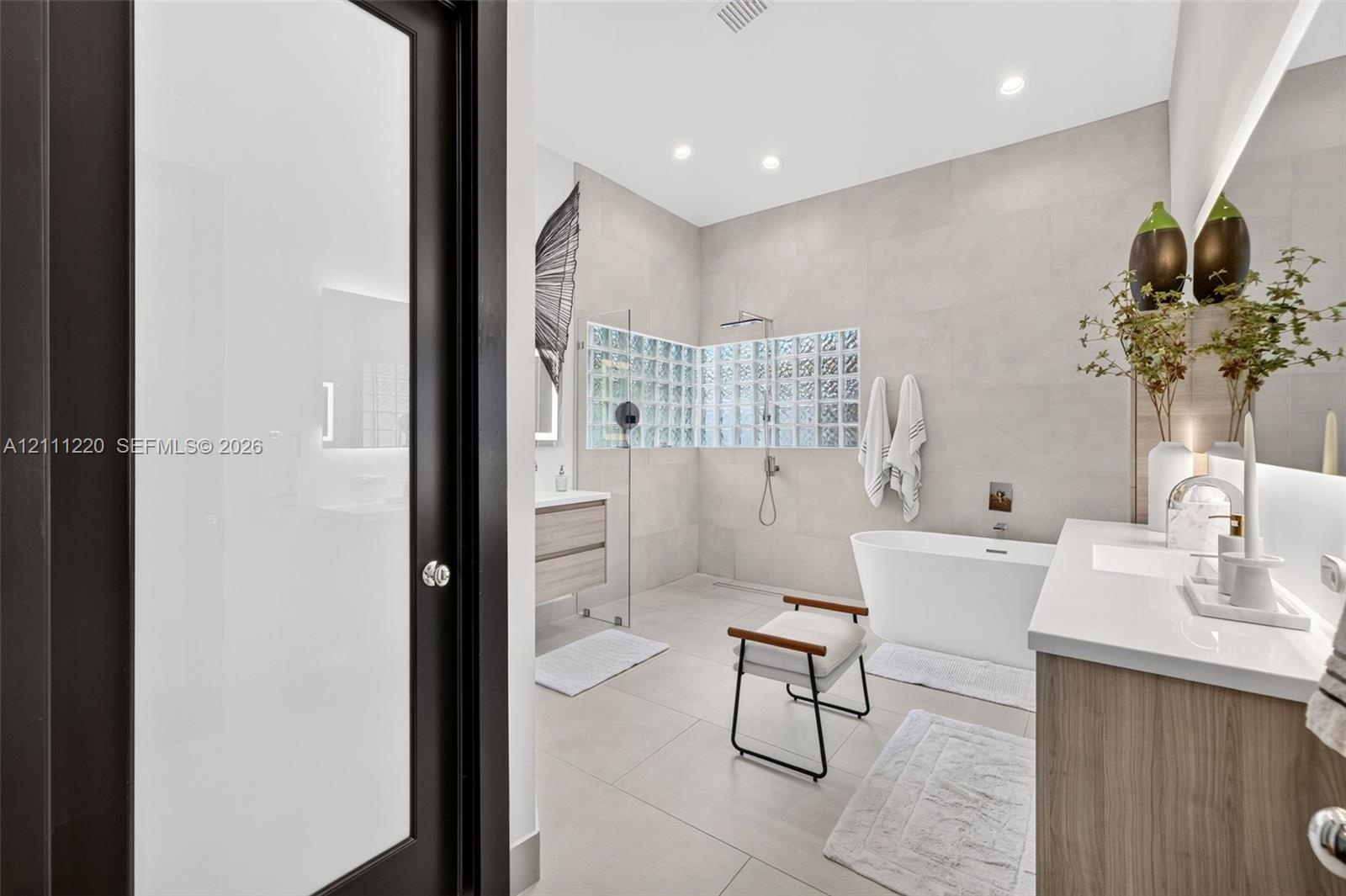 Bathroom featuring large-format tile flooring and walls, a freestanding soaking tub, a wet-room style shower enclosure with glass block wall detailing