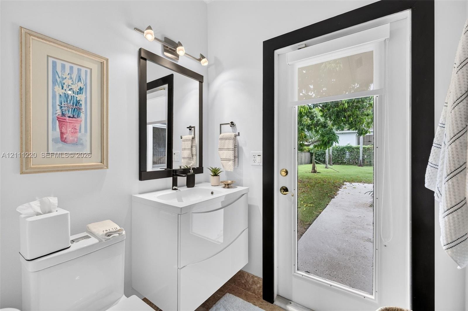 Modern floating vanity with a vessel sink, dark-framed vanity mirror, multi-bulb wall-mounted lighting, and an exterior glass door leading to a concre