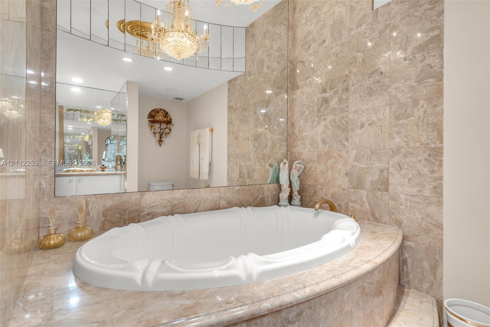 Oval soaking tub featuring a decorative basin, surrounded by large-format natural stone tiling with an expansive wall-to-wall mirror, crystal-style ch