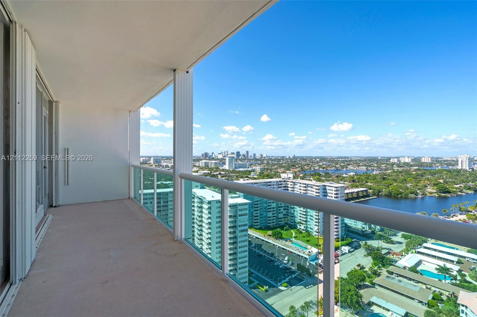 Covered balcony featuring concrete flooring, a glass-panel railing with a metallic handrail, and an expansive cityscape and waterway backdrop