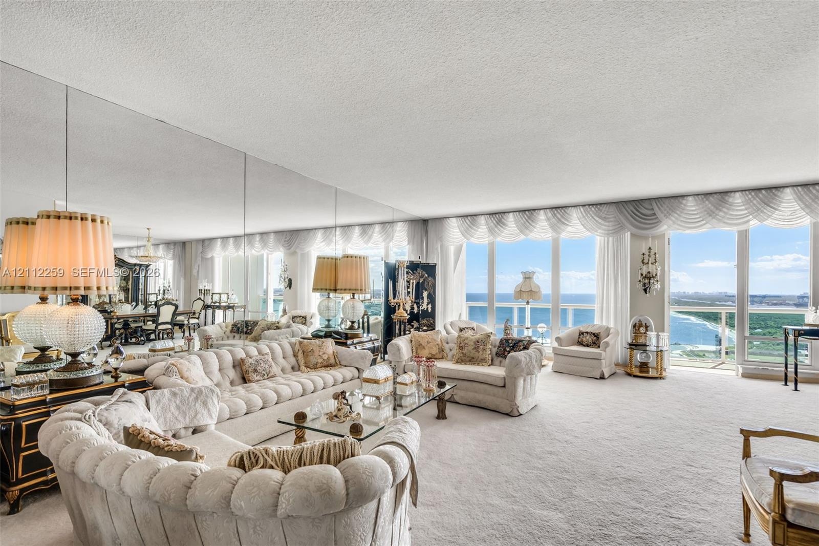 Expansive living area featuring floor-to-ceiling windows, wall-to-wall mirrored accent panels, and balcony access with coastal scenery