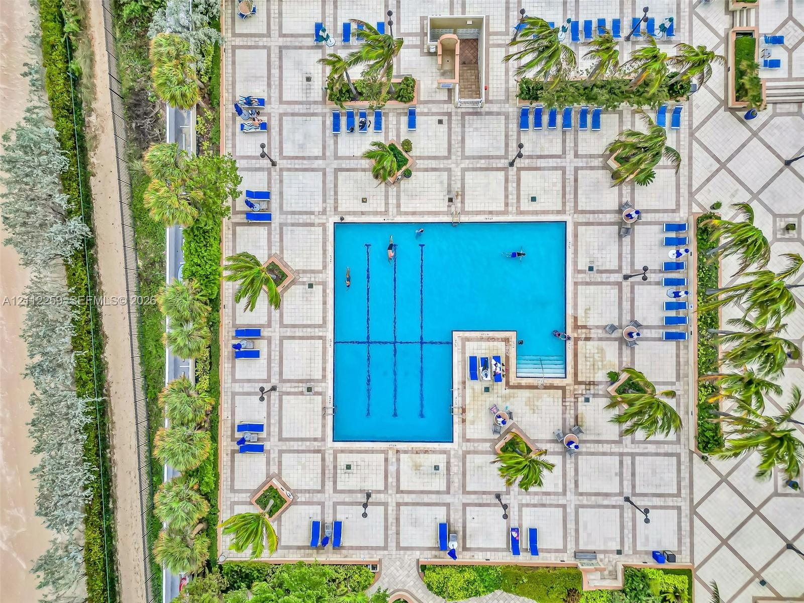 Geometric swimming pool with lap lanes and integrated stairs, surrounded by extensive patterned stone tiling, tropical landscaping, and mature palm tr