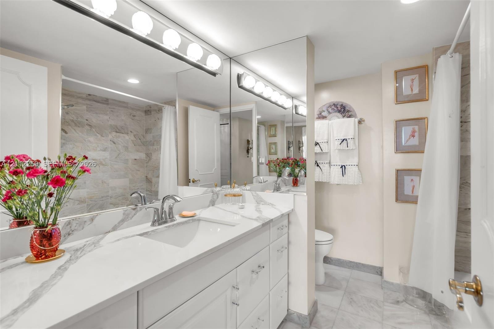 Bathroom featuring a white vanity with stone-patterned countertop, integrated basin, full-width mirror, multi-bulb light fixture, and stone-tiled show