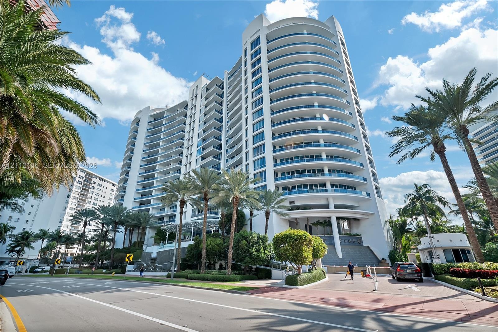 High-rise residential tower featuring a curved white facade, continuous glass-railed balconies, and a paved gated entrance