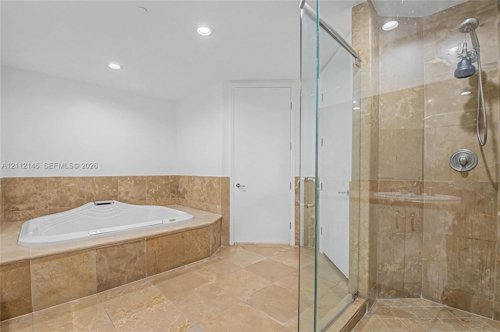 Soaking tub with a natural stone tile surround, glass-enclosed shower, polished metal fixtures, and stone tile flooring
