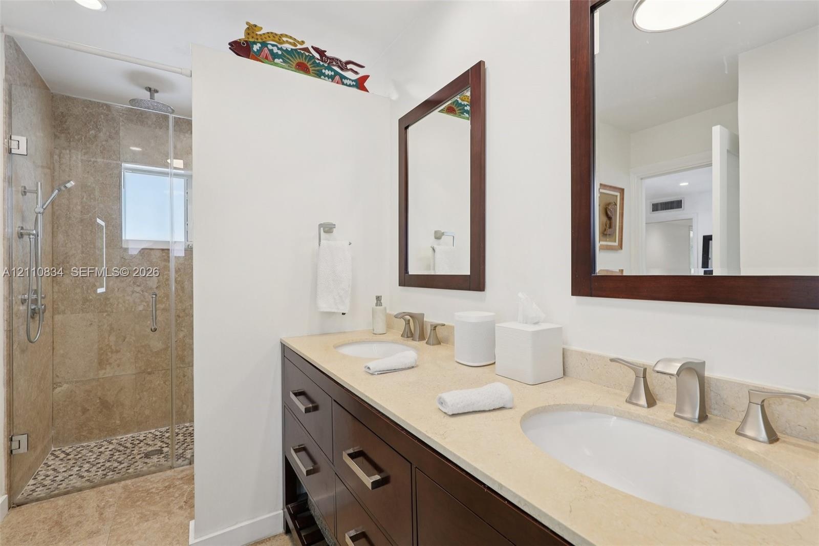 Double vanity with stone countertop, dark wood-finish cabinetry, frameless glass shower enclosure, stone-tiled shower wall, and integrated shower rain