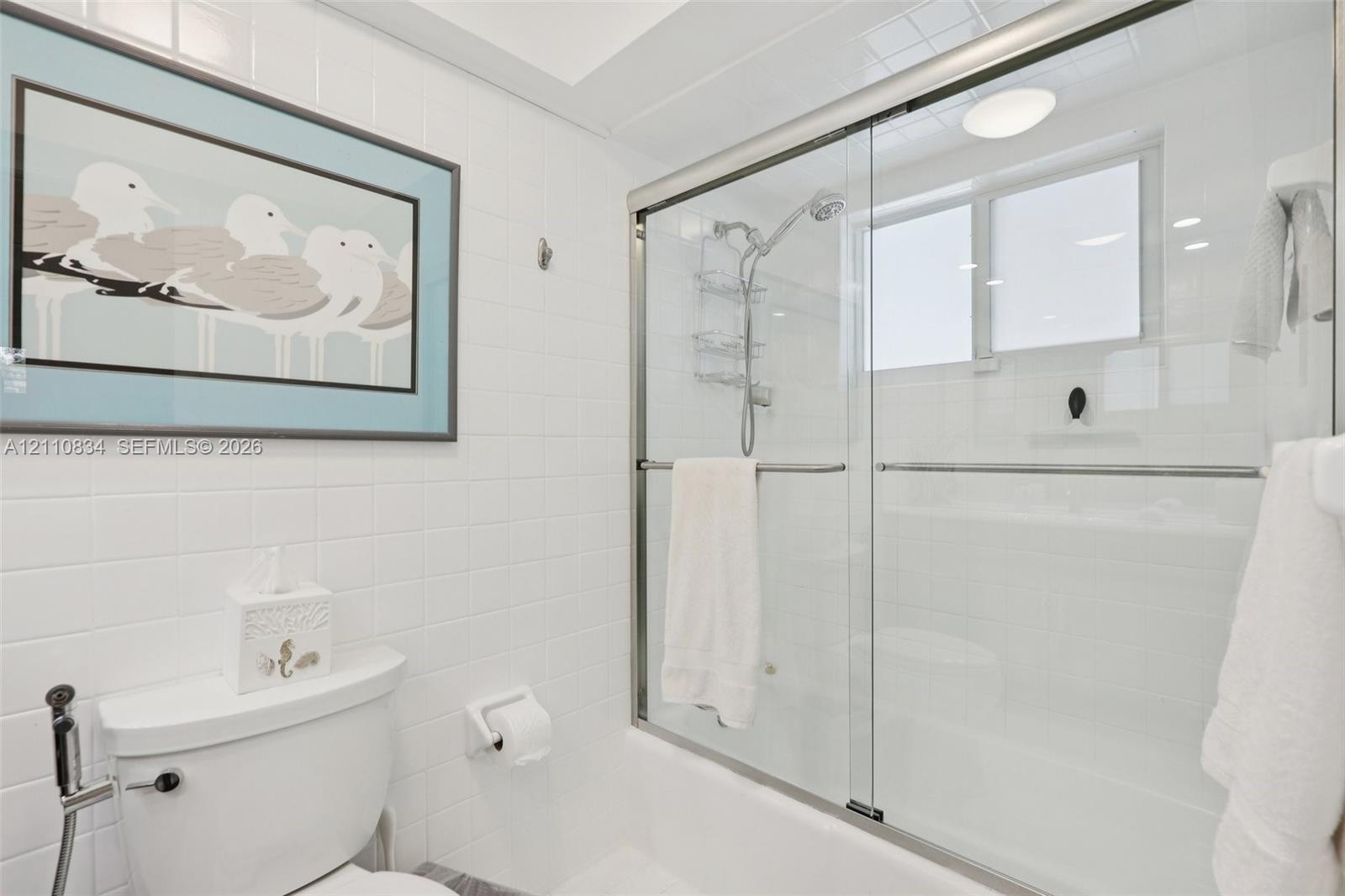 Bathroom featuring white tile wall surround, glass-enclosed shower-tub combination, chrome plumbing fixtures, and a recessed ceiling light