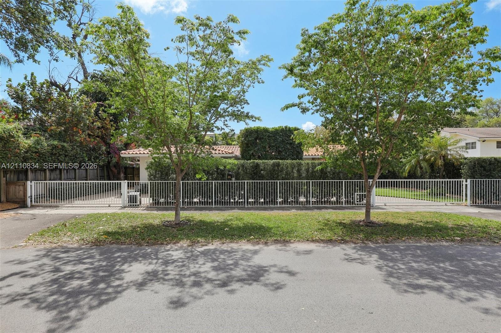 White metal perimeter fencing, gated driveway access, mature landscaping, and established trees