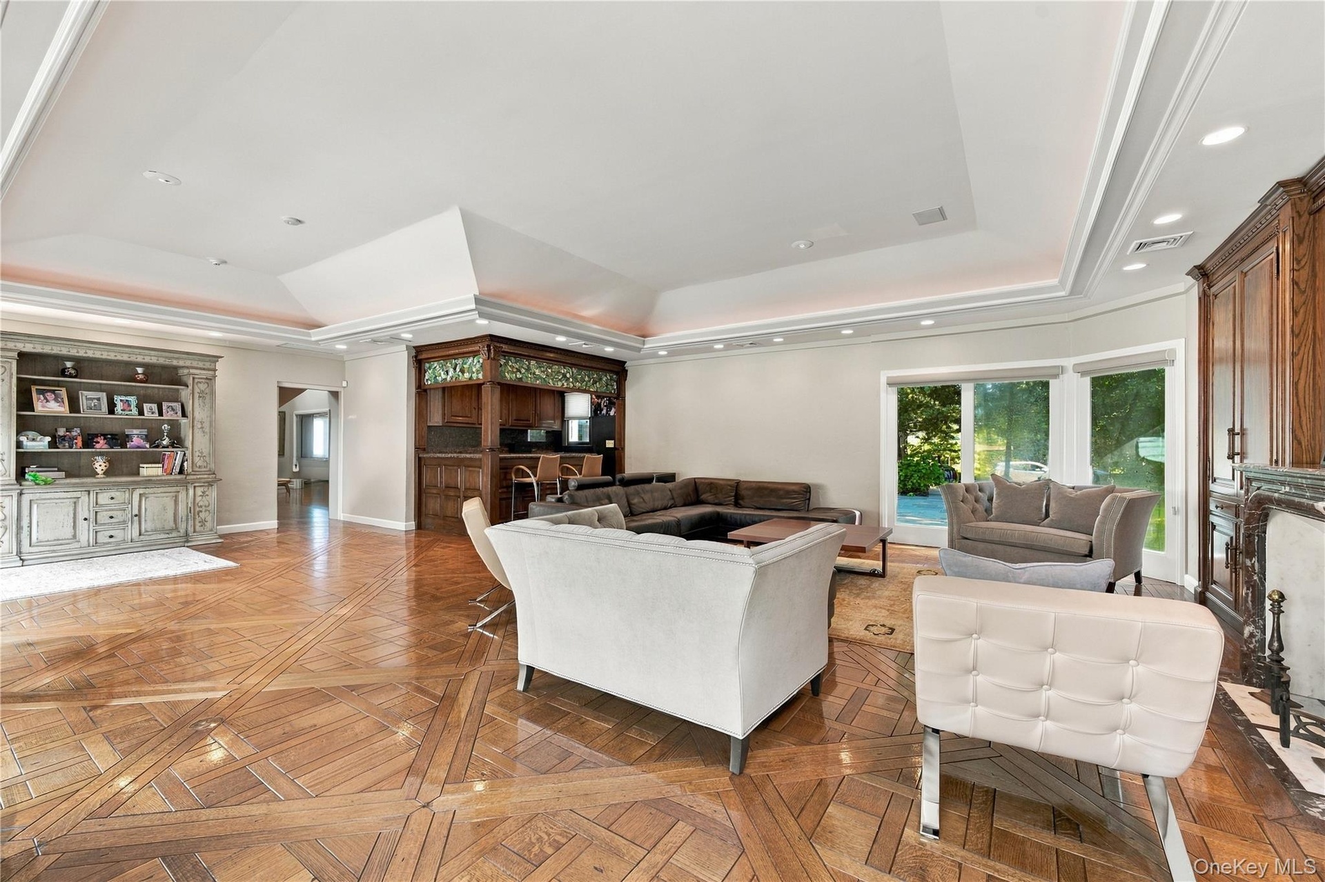 Expansive room featuring intricate parquet wood-finish flooring, a tiered tray ceiling with recessed lighting, custom built-in cabinetry, a wet bar wi